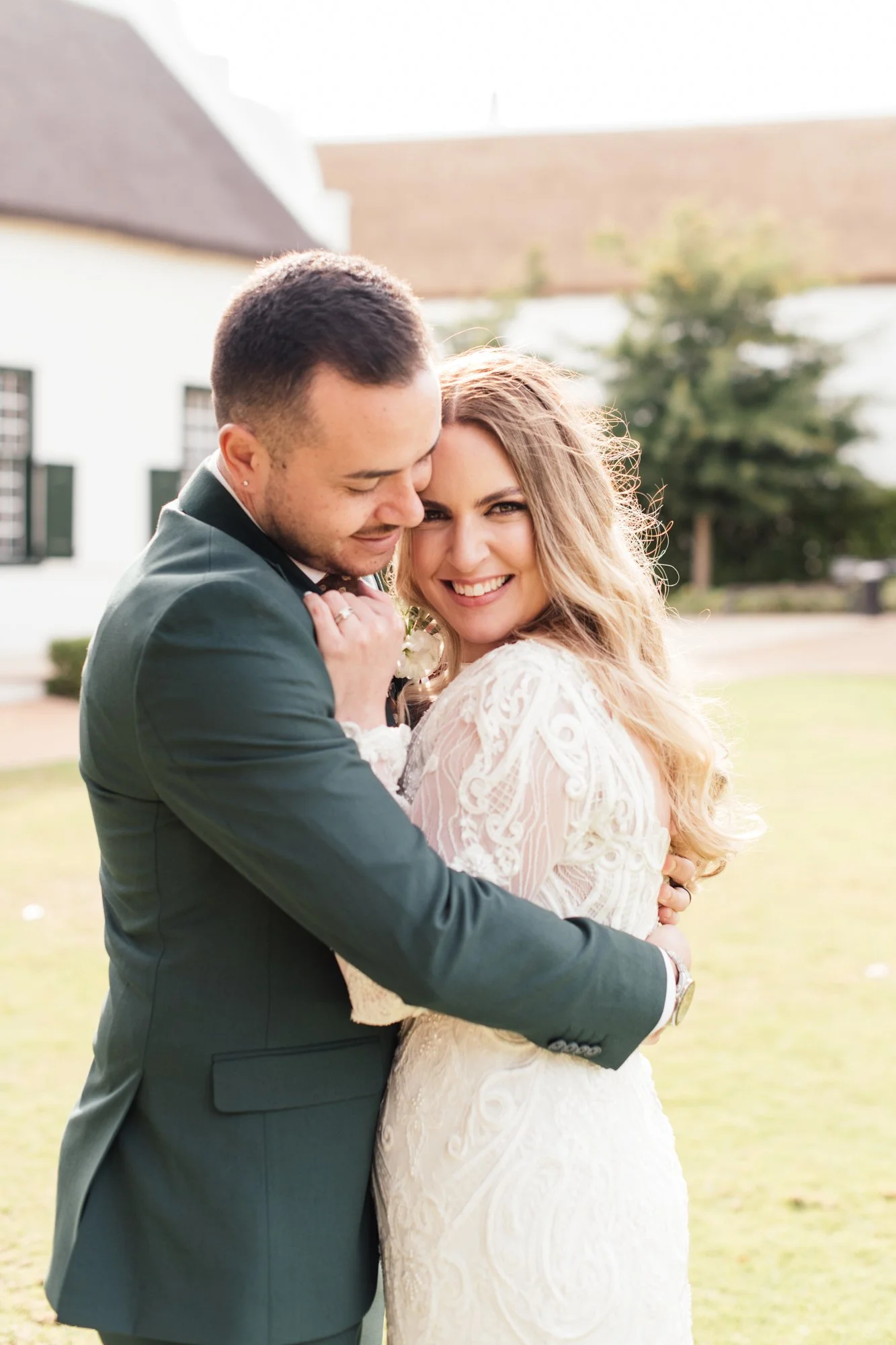 A happy couple in wedding attire embracing outdoors, smiling and looking at the camera.