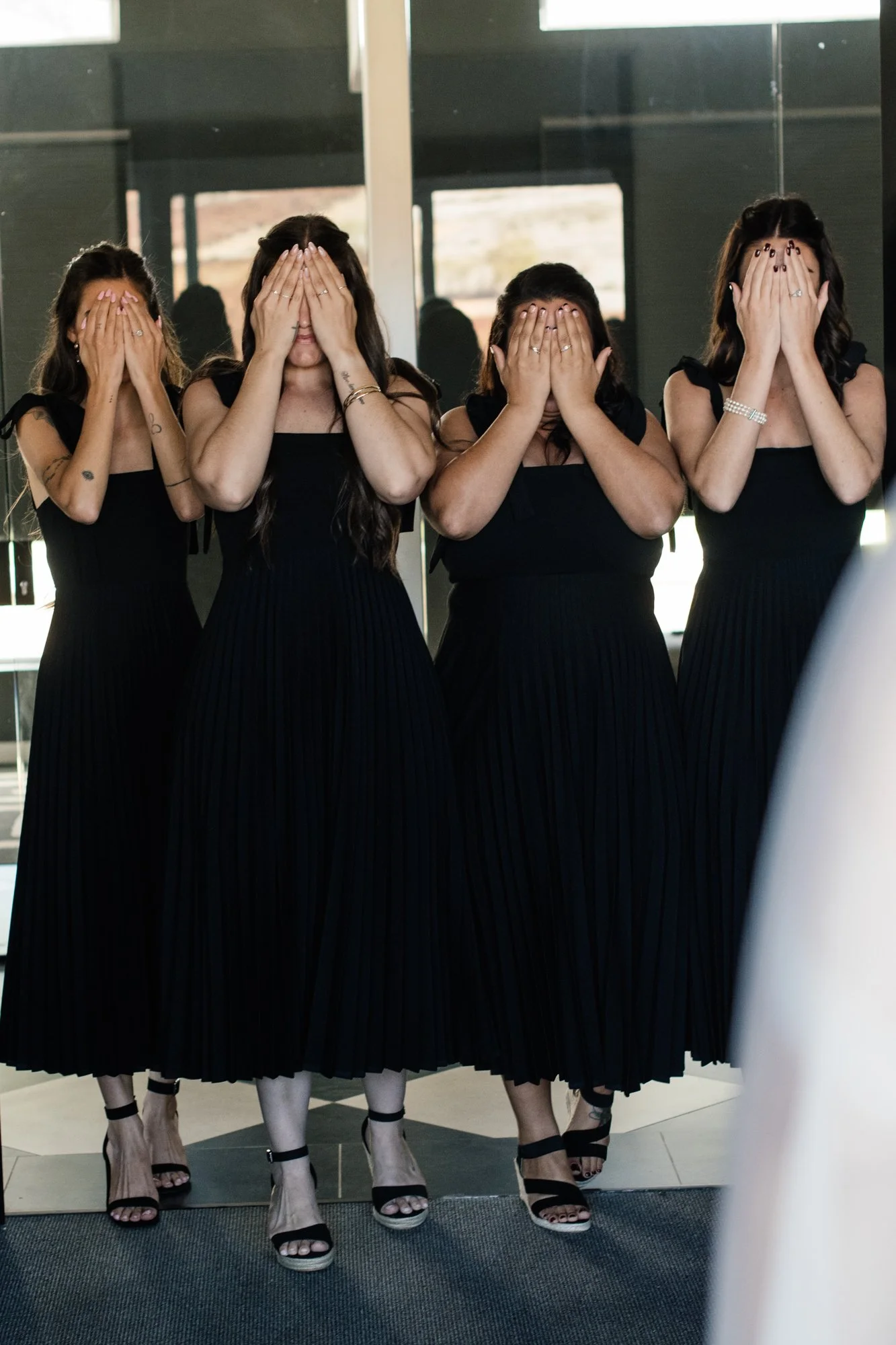Four women in black dresses and high-heeled shoes stand in front of a mirror, covering their faces with their hands.