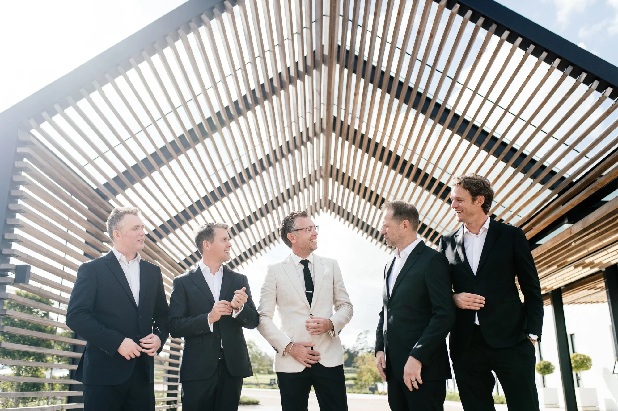 A group of groomsman with the groom in formal suits standing outdoors and engaging in conversation, underneath a modern architectural structure with wooden slats.