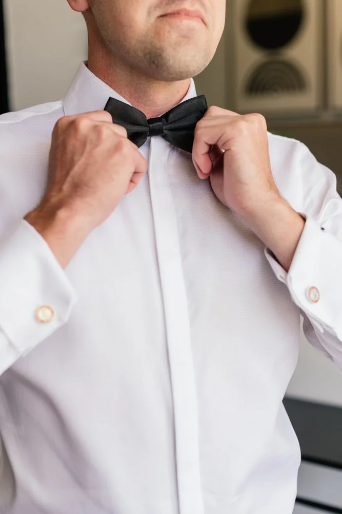 Close-up of a man adjusting a black bow tie while wearing a white dress shirt.