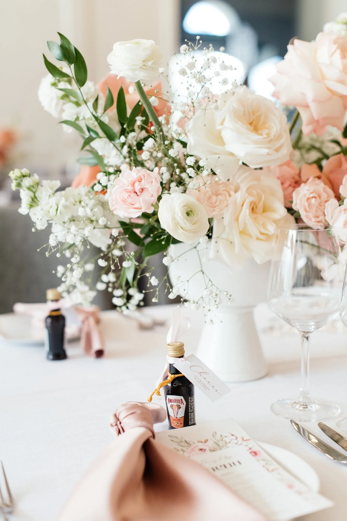Close-up of a flower arrangement of white, pink, and peach roses with baby's breath and green leaves on a table at a celebration or wedding reception.