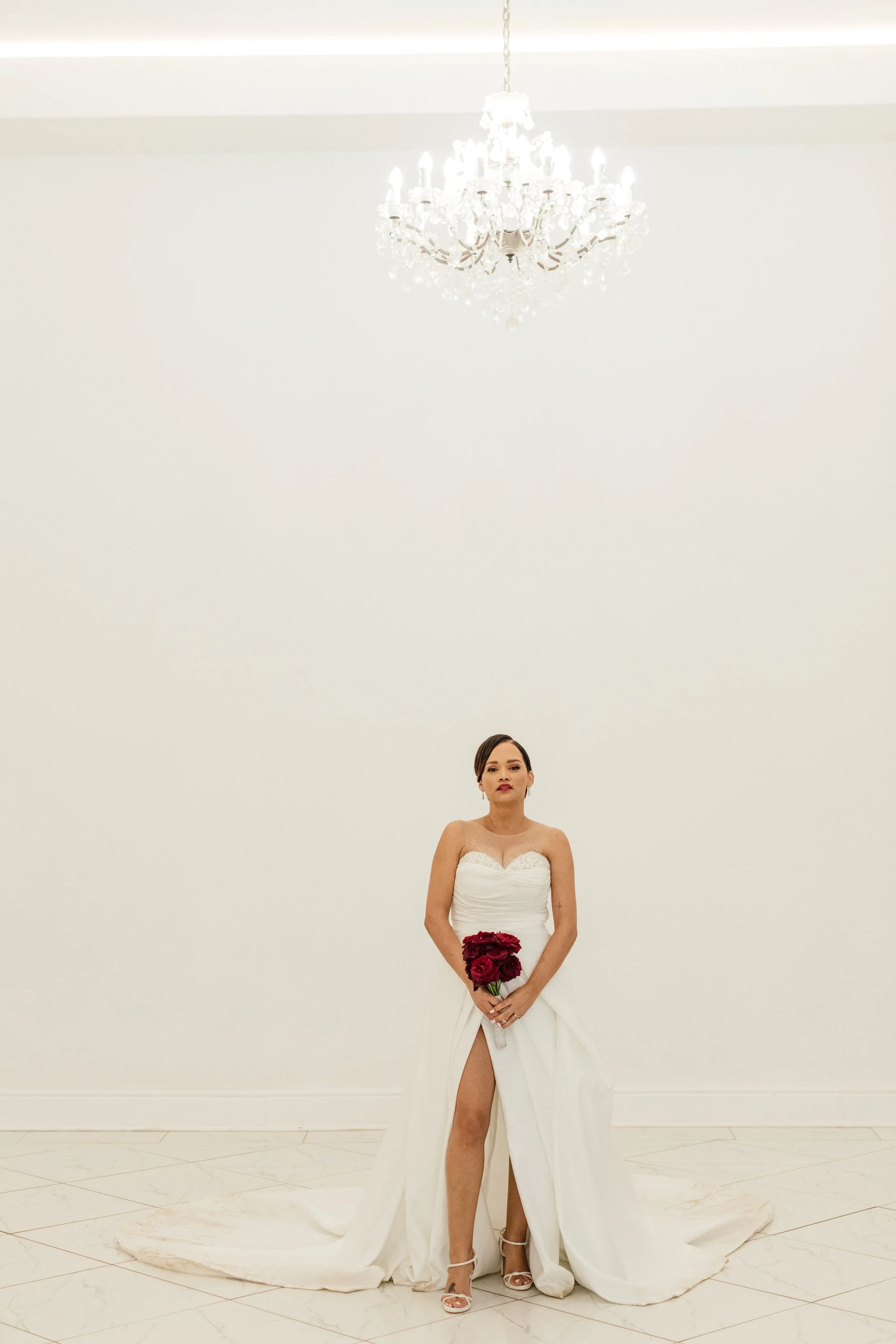 A woman in a white wedding dress with a high slit, holding a bouquet of red roses, standing in a minimalistic white room with a large chandelier overhead.