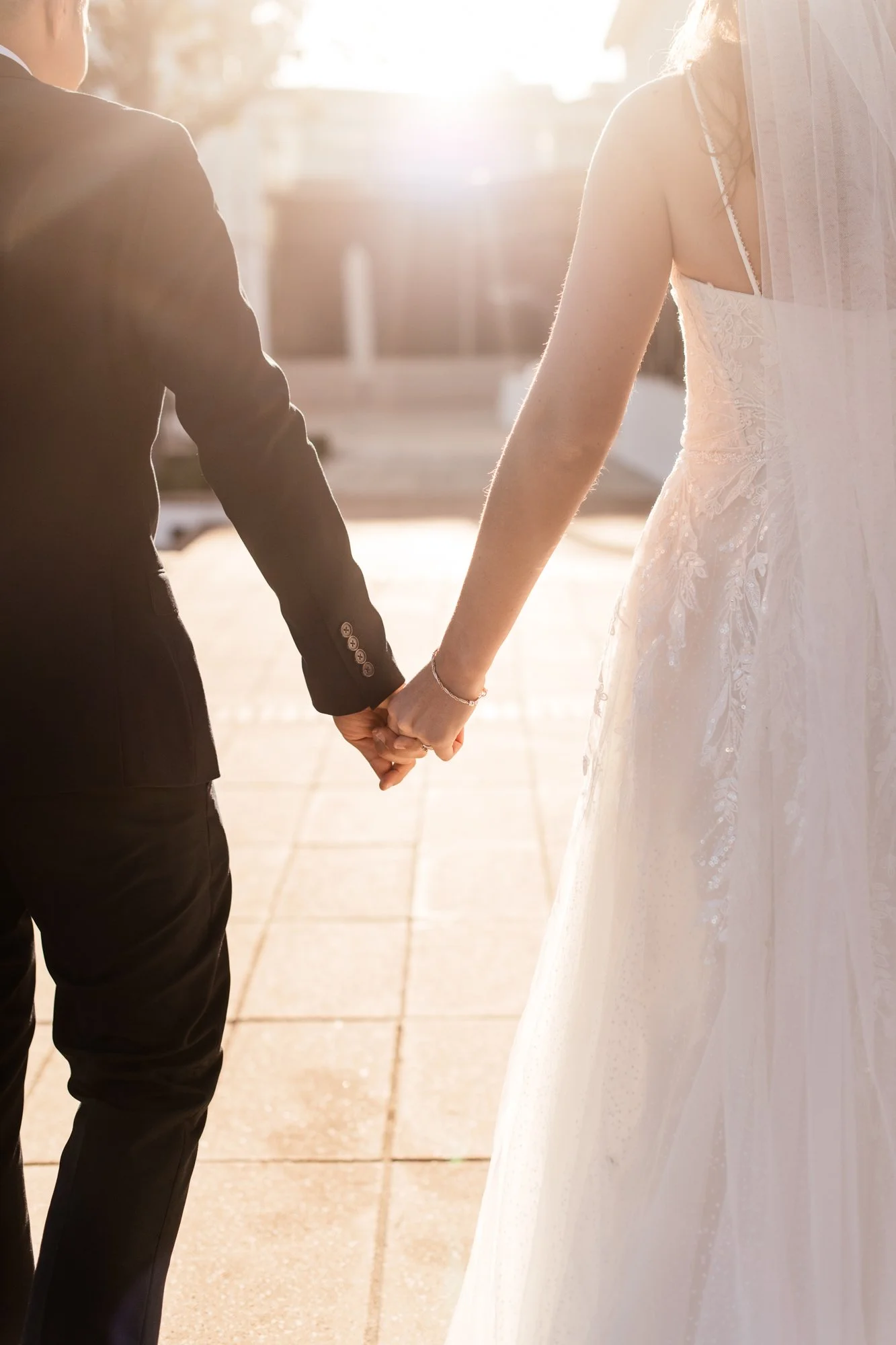 A couple holding hands, with the man wearing a black suit and the woman wearing a white lace wedding dress, standing outdoors in sunlight.