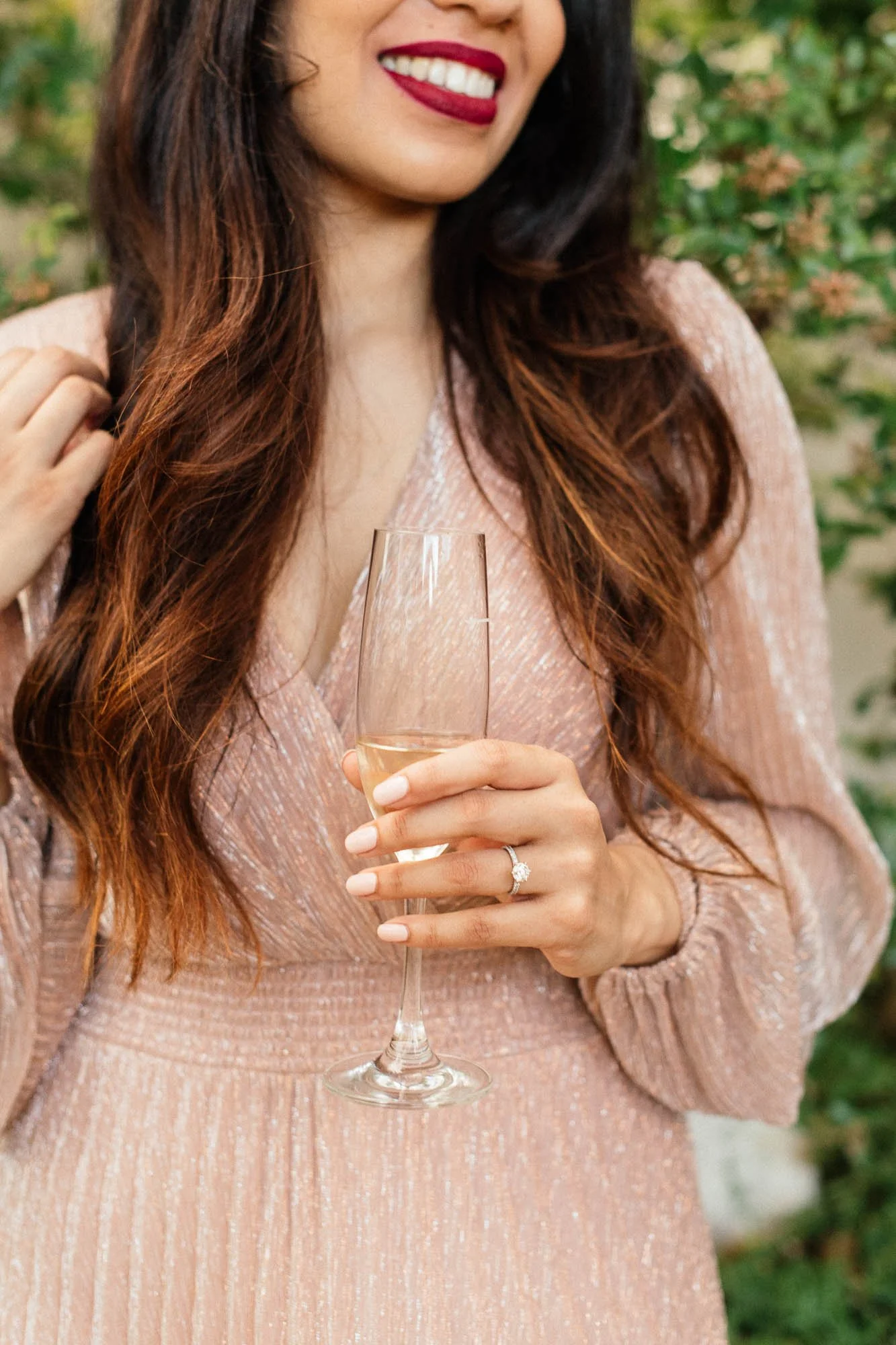 A woman with long brown hair wearing a pink, shimmery dress, holding a glass of white wine, and wearing a ring with a large gemstone.