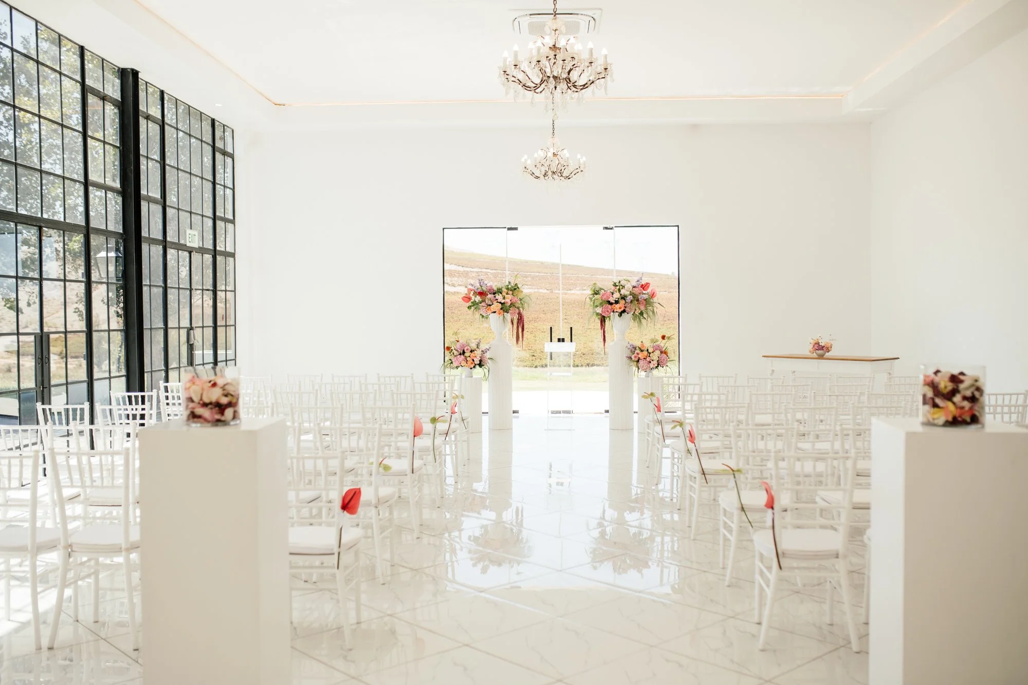 Elegant wedding ceremony setup in a bright, modern venue with white chairs, tall floral arrangements, crystal chandeliers, and large windows revealing a scenic landscape outside.