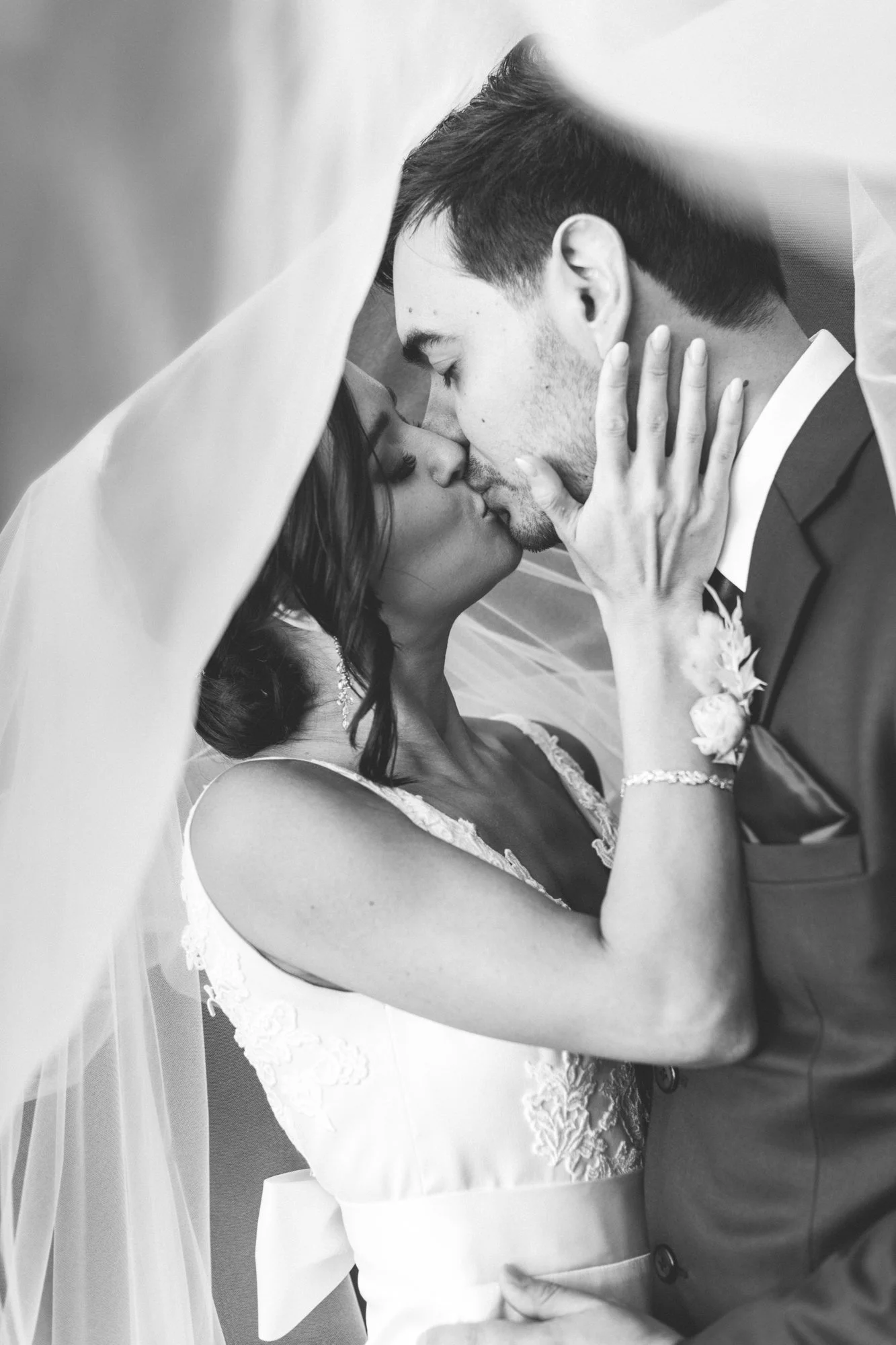 A black and white photo of a bride and groom kissing under a veil, with the bride’s hand on the groom's face.