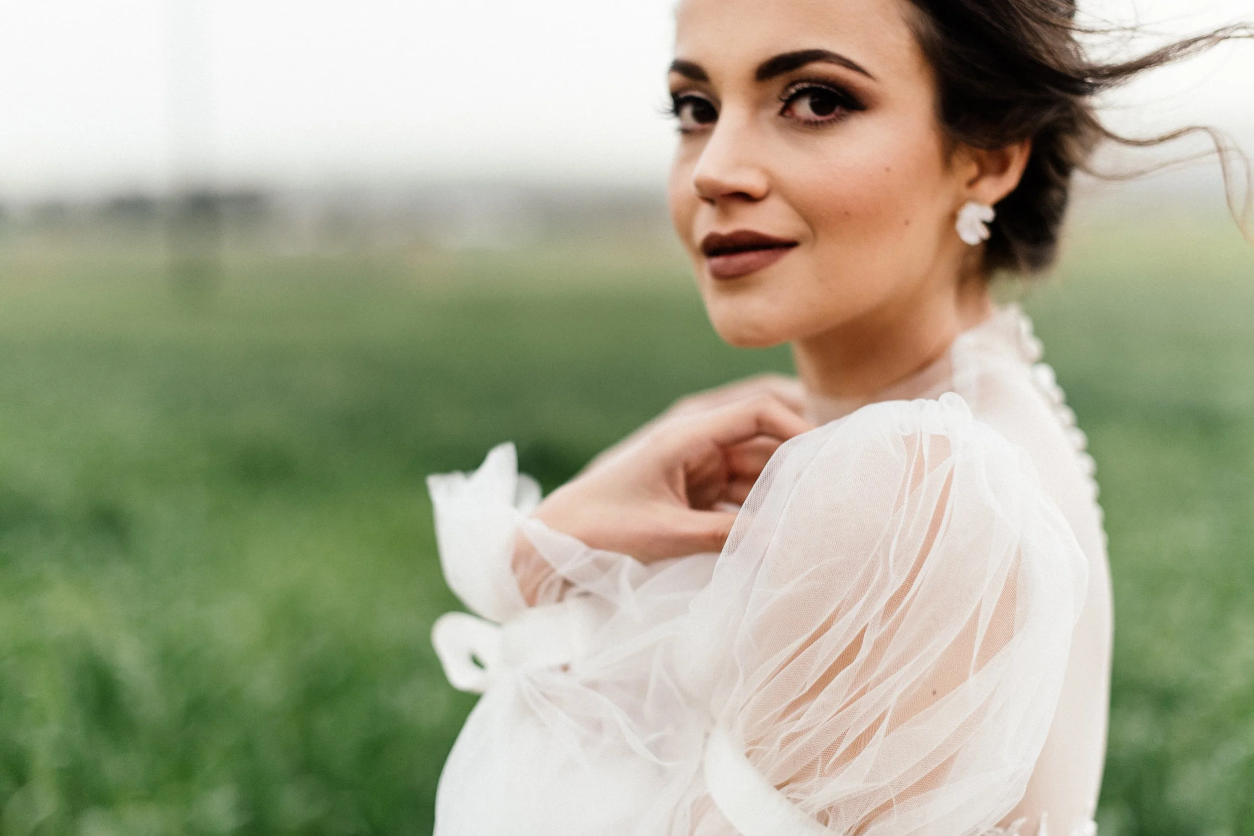 A woman with dark hair and earrings, wearing a sheer white dress, standing outdoors in a green field, looking over her shoulder.