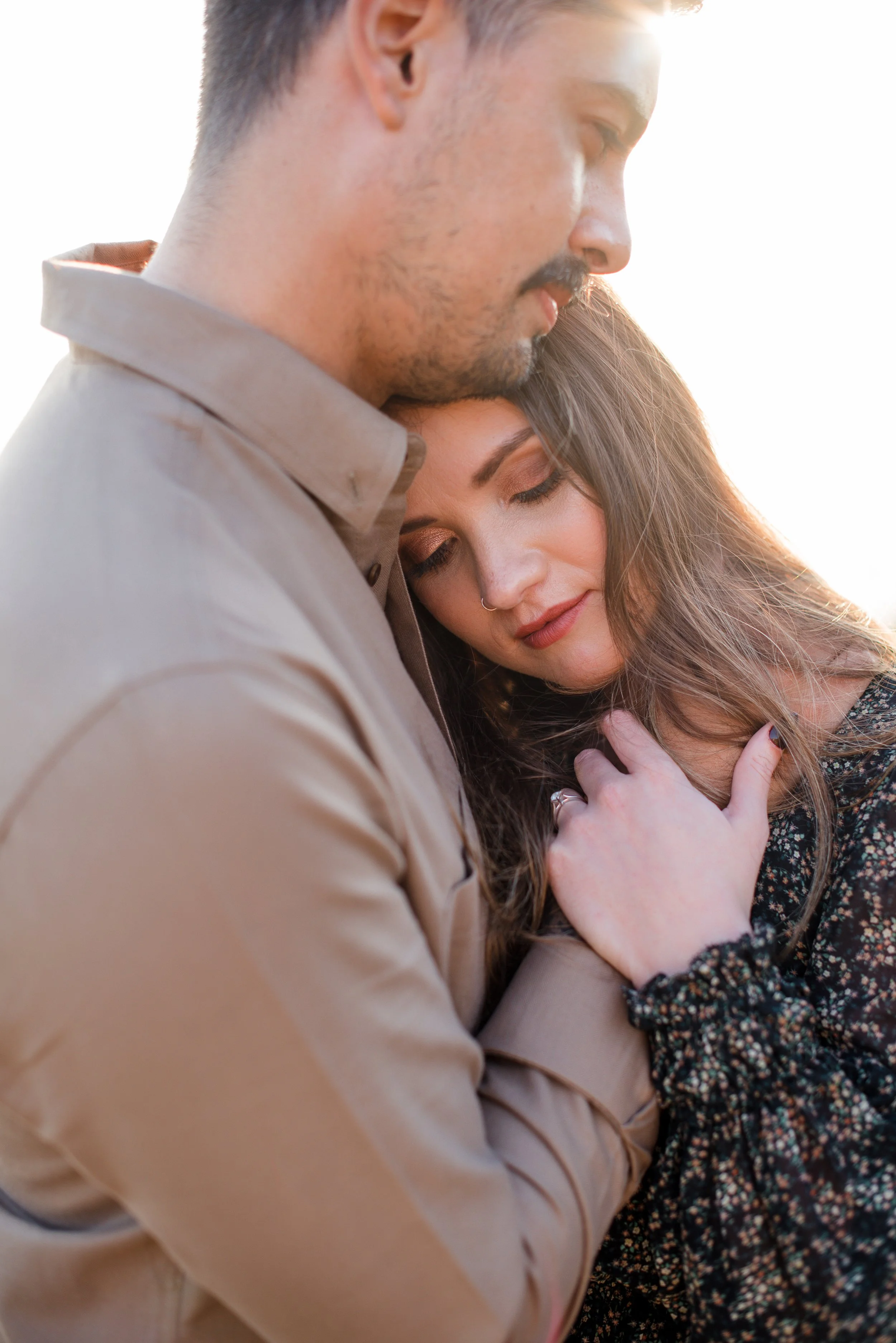 A man with light skin, dark hair, and a beard hugs a woman with light skin and long brown hair, both with their eyes closed, outdoors with bright sunlight.