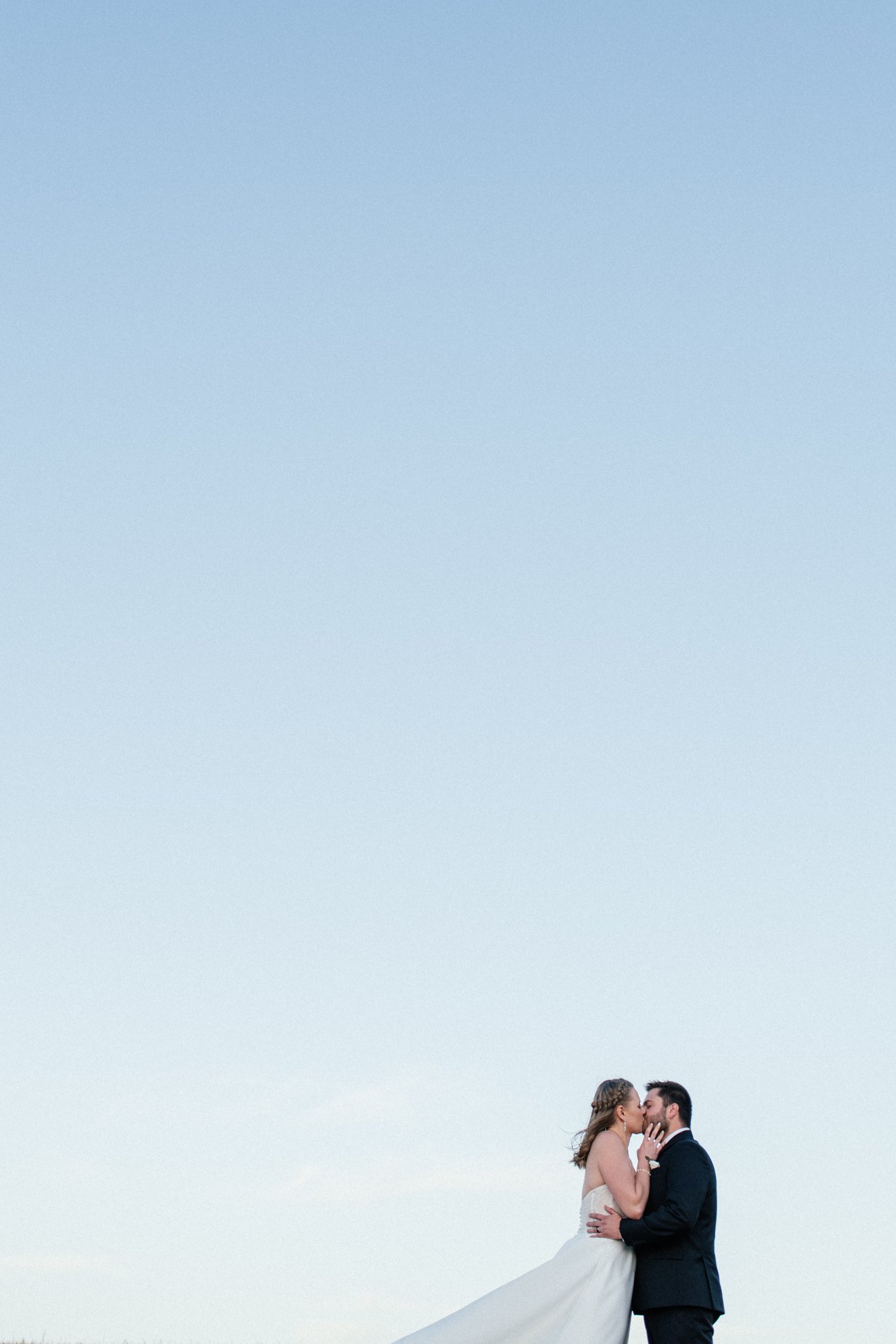 A bride and groom kissing outdoors, with an expansive sky overhead.