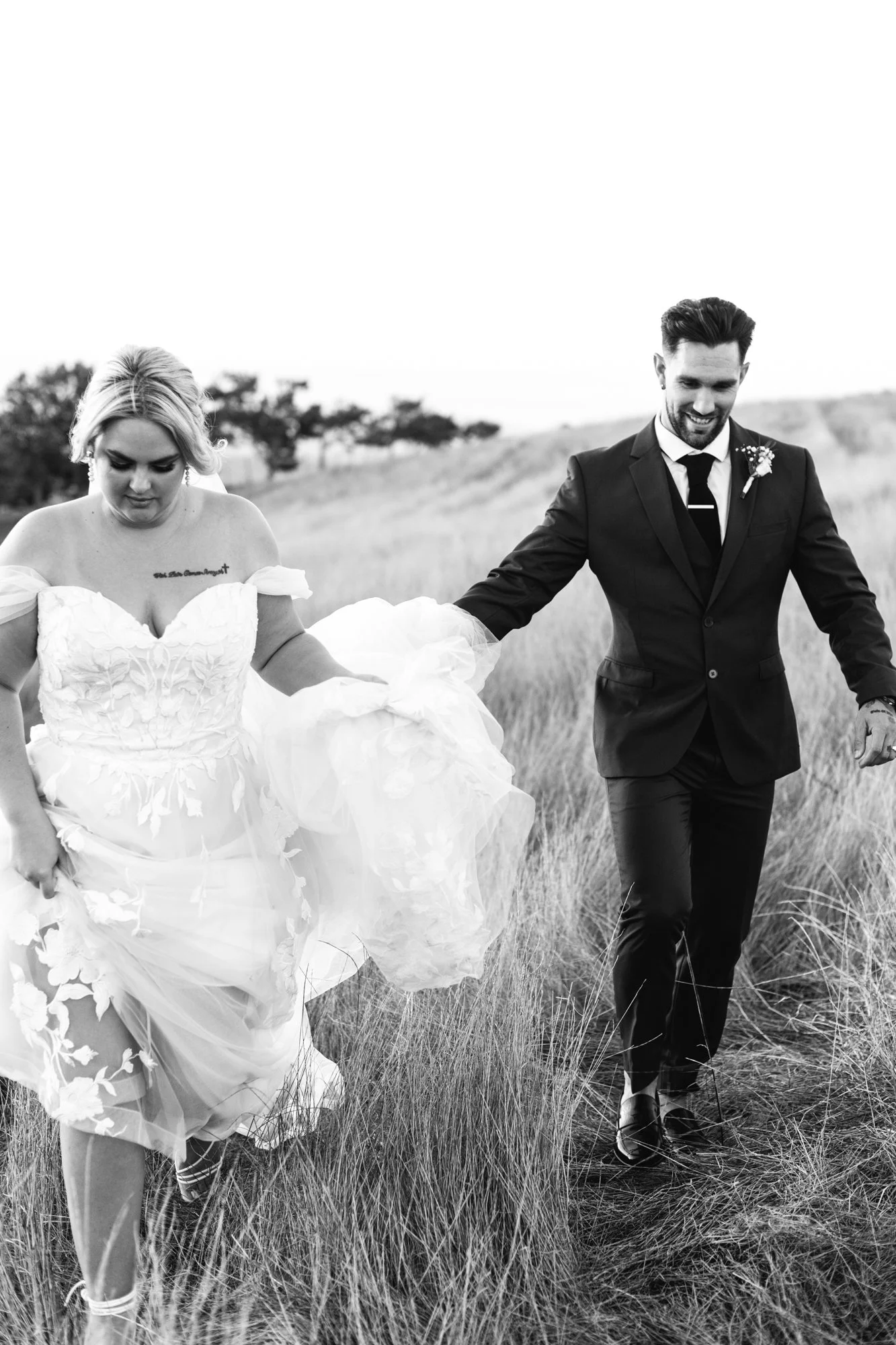 A bride and groom walking hand in hand through a grassy field.