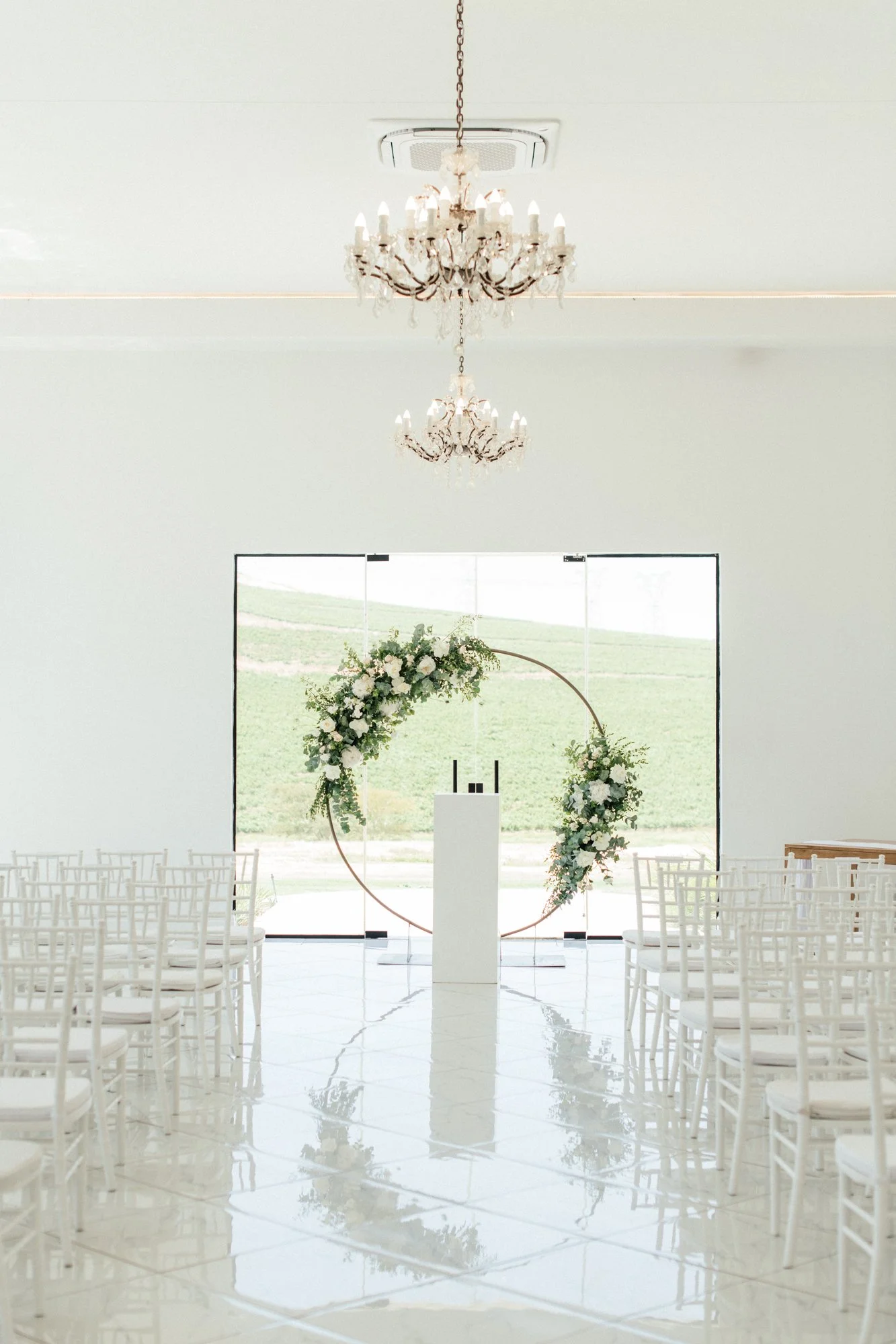 A modern wedding ceremony setup inside with white chairs, a large circular floral arch, and a minimalist white podium, with chandeliers hanging from the ceiling and natural light coming through the glass doors.