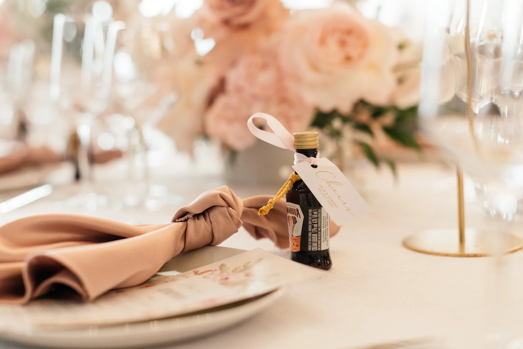 Close-up of a table setting at a celebration, featuring a small dark bottled drink with a white ribbon and tag, a pink napkin, and blurred floral arrangements and glassware in the background.