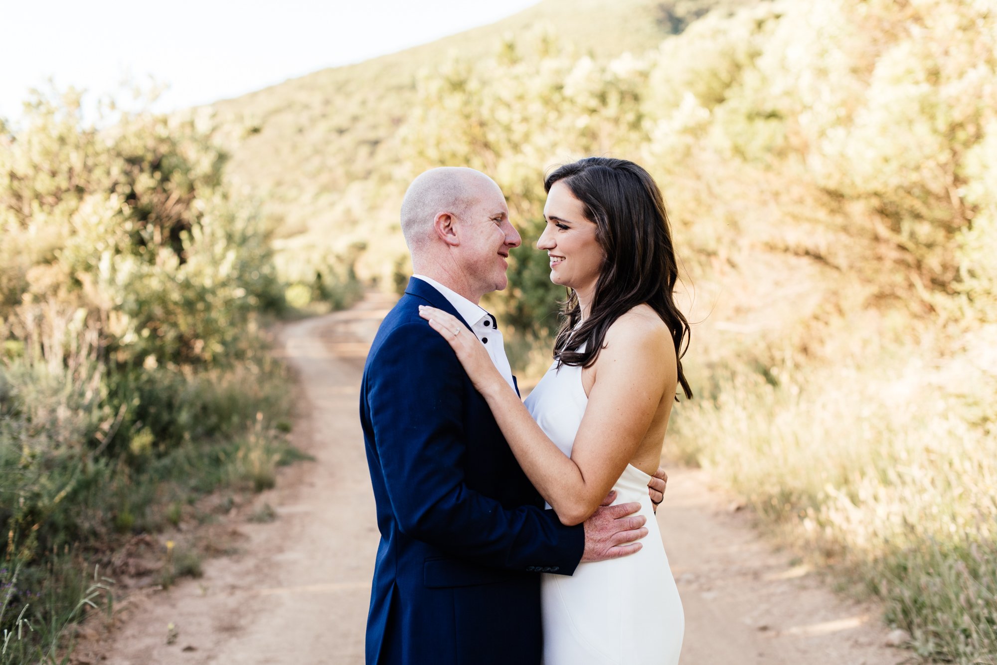 A couple dressed in formal attire, standing outdoors on a dirt path surrounded by trees and hills, smiling at each other closely.