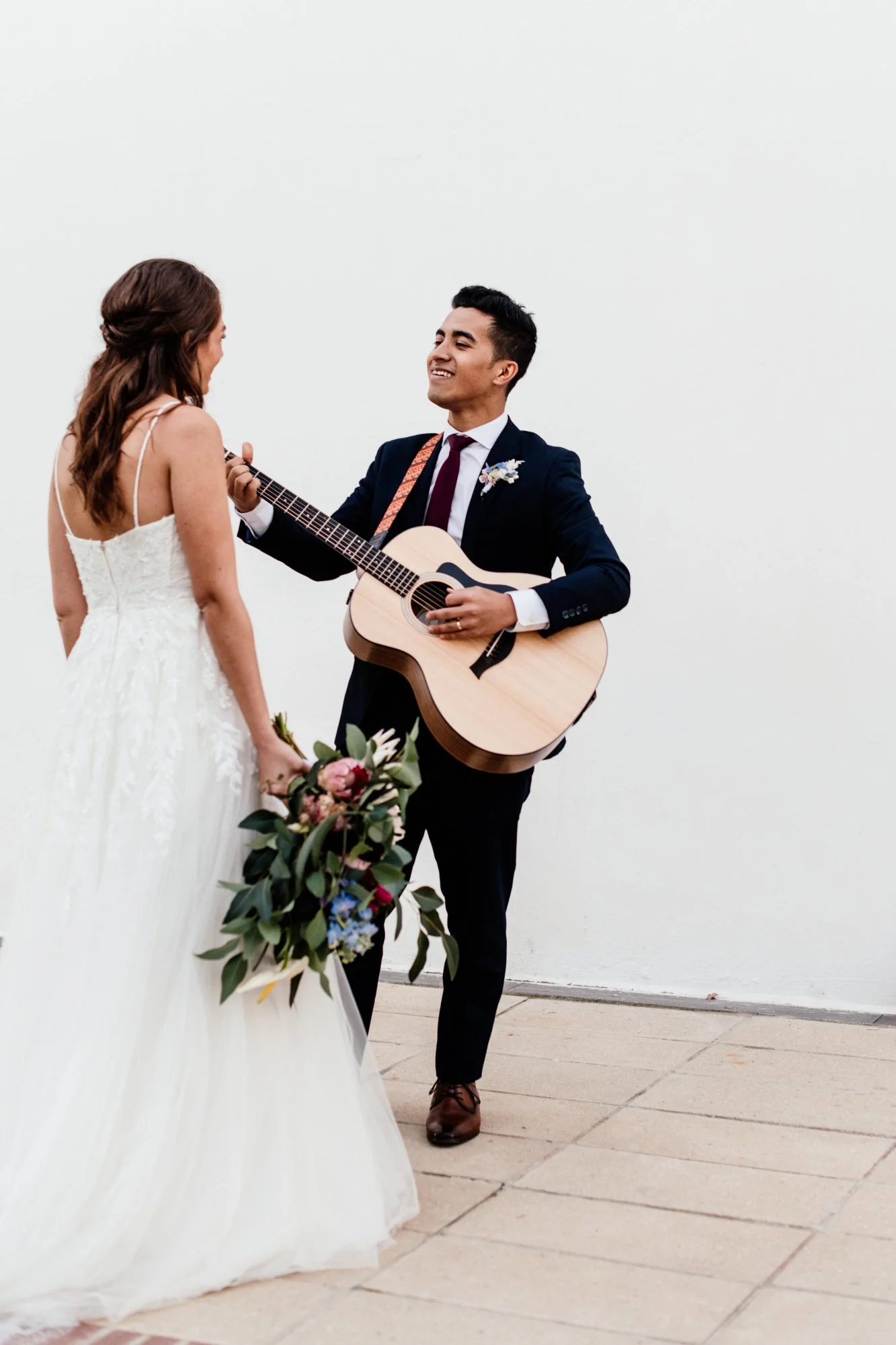 A bride and groom exchange vows during a wedding ceremony. The groom is playing an acoustic guitar, and the bride holds a bouquet of flowers. They stand against a plain white wall.