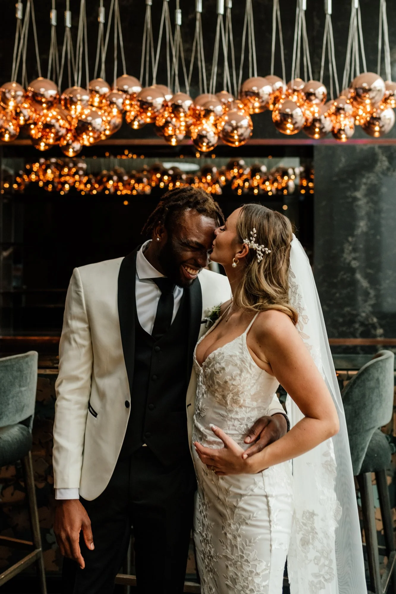 A bride and groom are dancing closely at their wedding reception indoors, surrounded by stylish decor including a mirror and hanging spherical light fixtures.