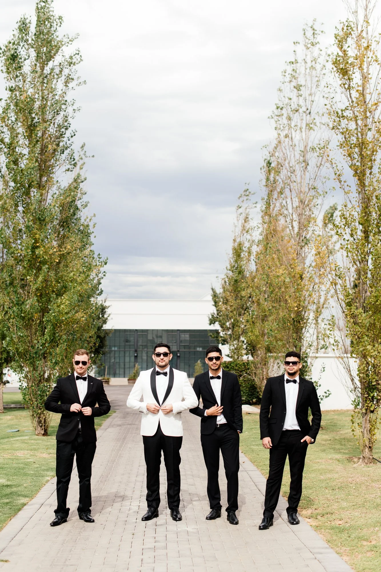 Five men dressed in tuxedos walking on a paved pathway outdoors during daytime, with trees and a modern building in the background.
