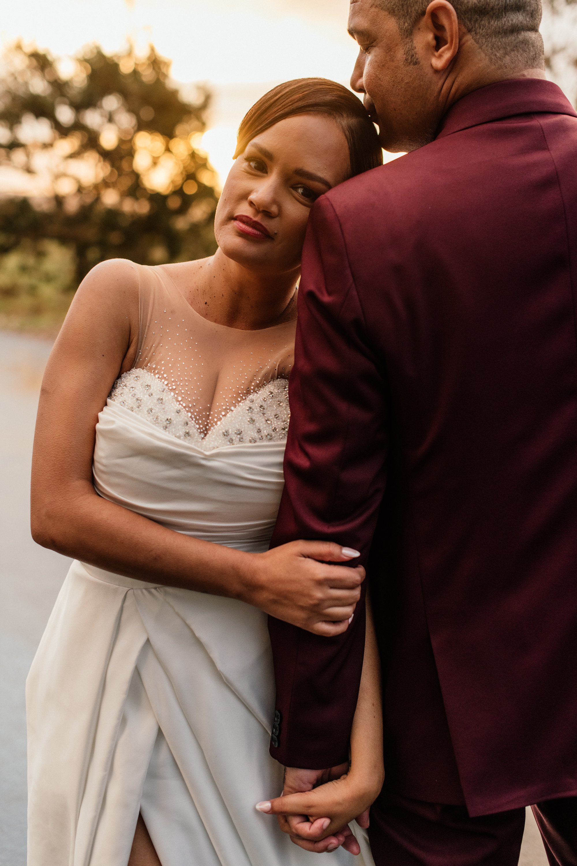 A woman in a wedding dress and a man in a maroon suit holding hands, with their heads touching, during sunset outdoors.