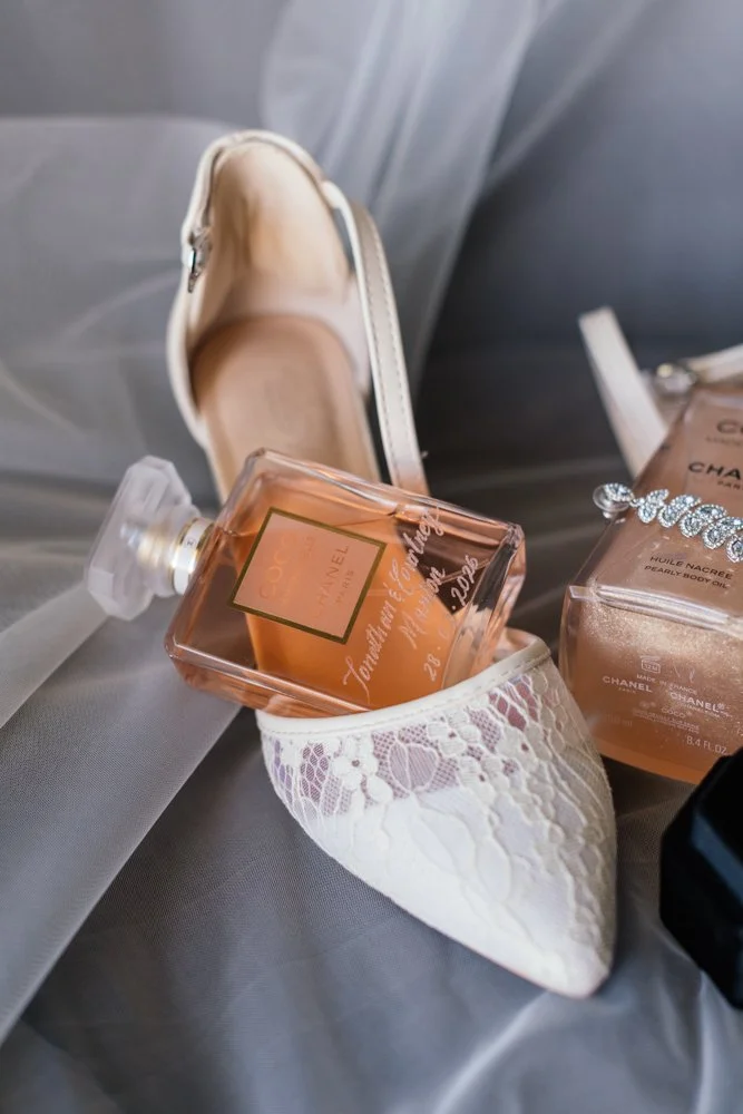 A white lace flat shoe containing a bottle of Coach perfume, surrounded by a Chanel body oil, a black object, and decorated with a rhinestone bracelet.
