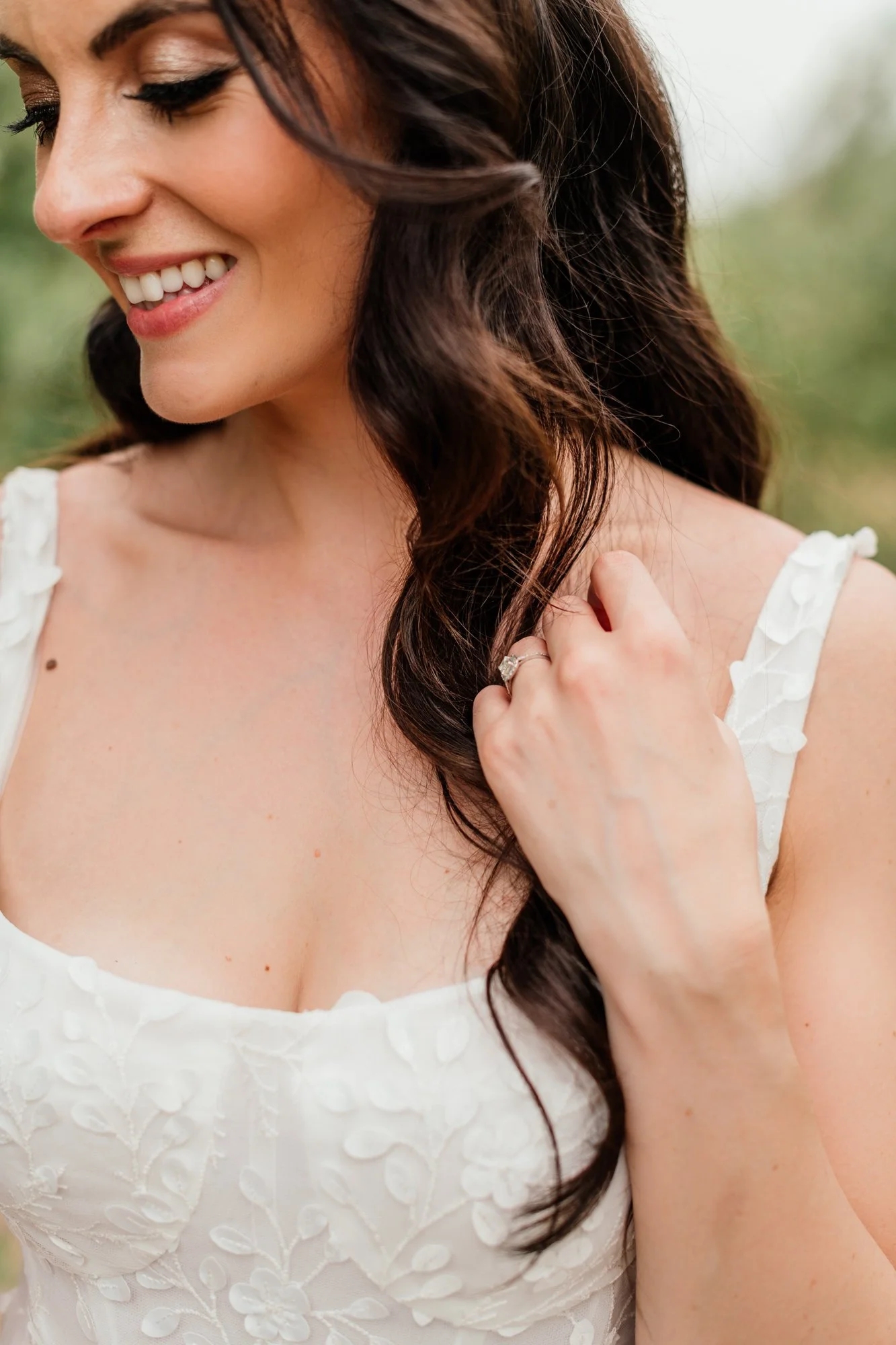 A woman with long, dark brown hair and light skin, smiling and looking down slightly, dressed in a white dress with embroidered floral details, outdoors with blurred greenery in the background.
