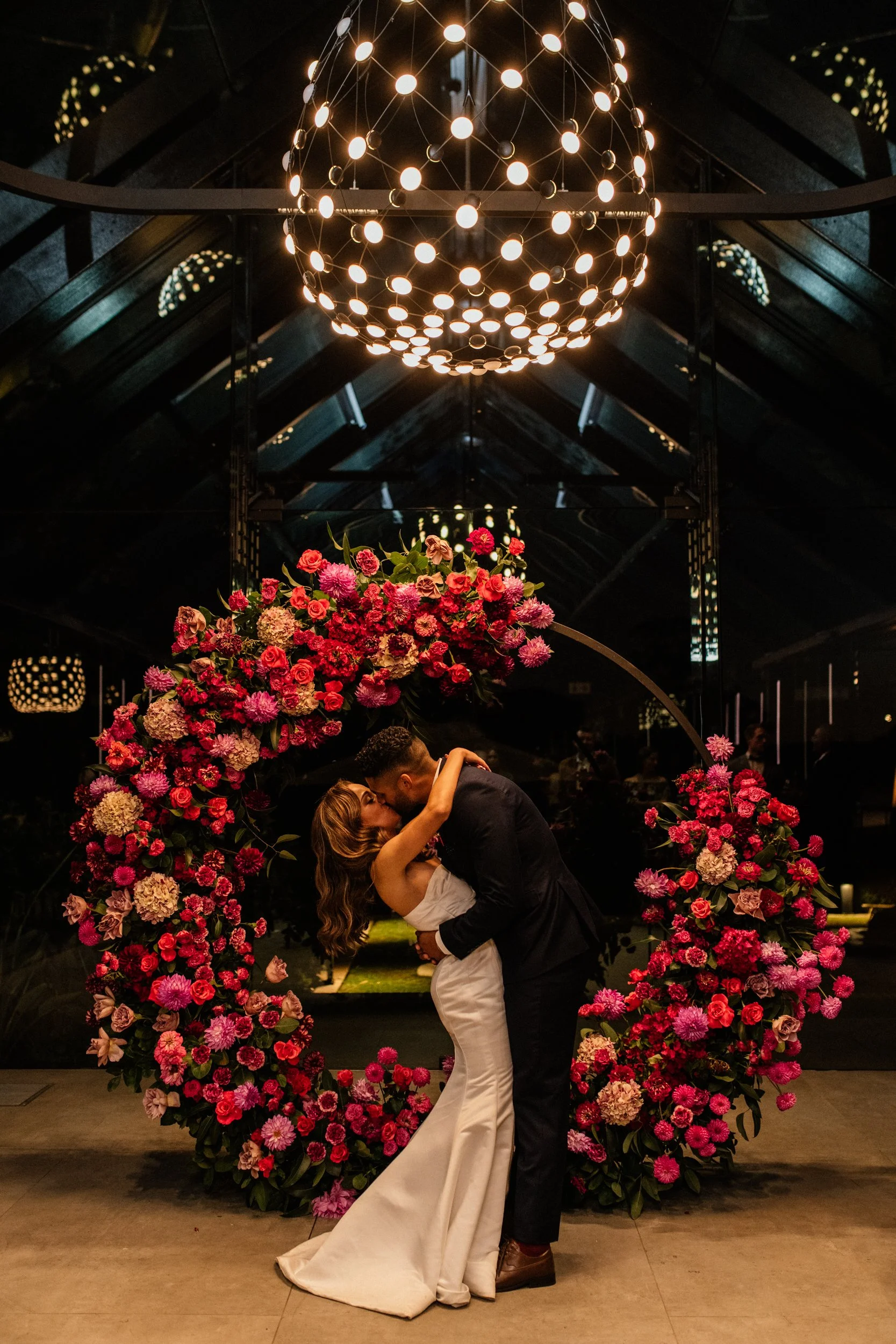 A couple sharing a kiss during their wedding, standing under a floral arch with pink and purple flowers, in a venue with a decorative chandelier overhead.