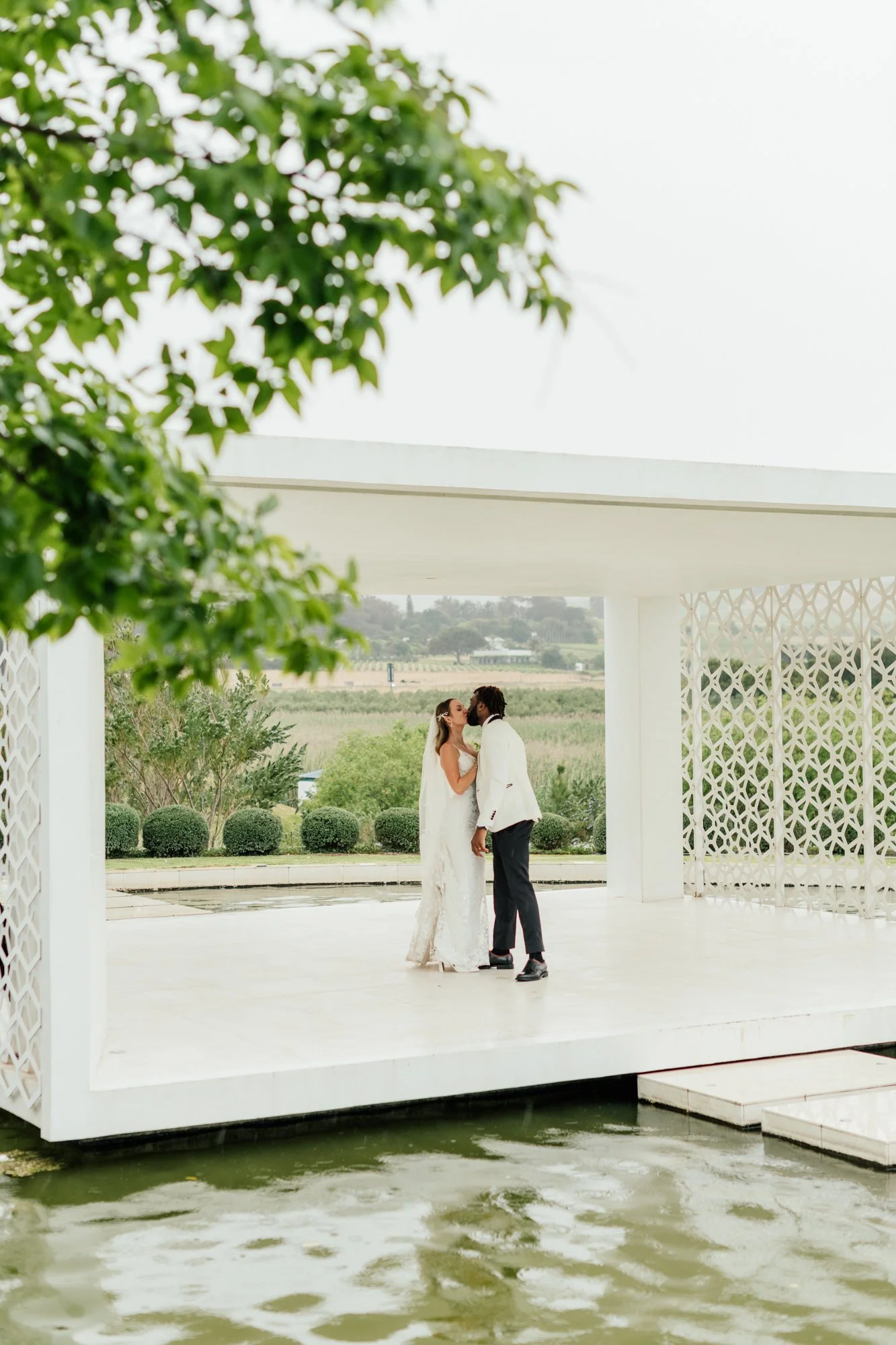 A bride and groom sharing a kiss on a modern white pavilion with water in the foreground and green landscape in the background.