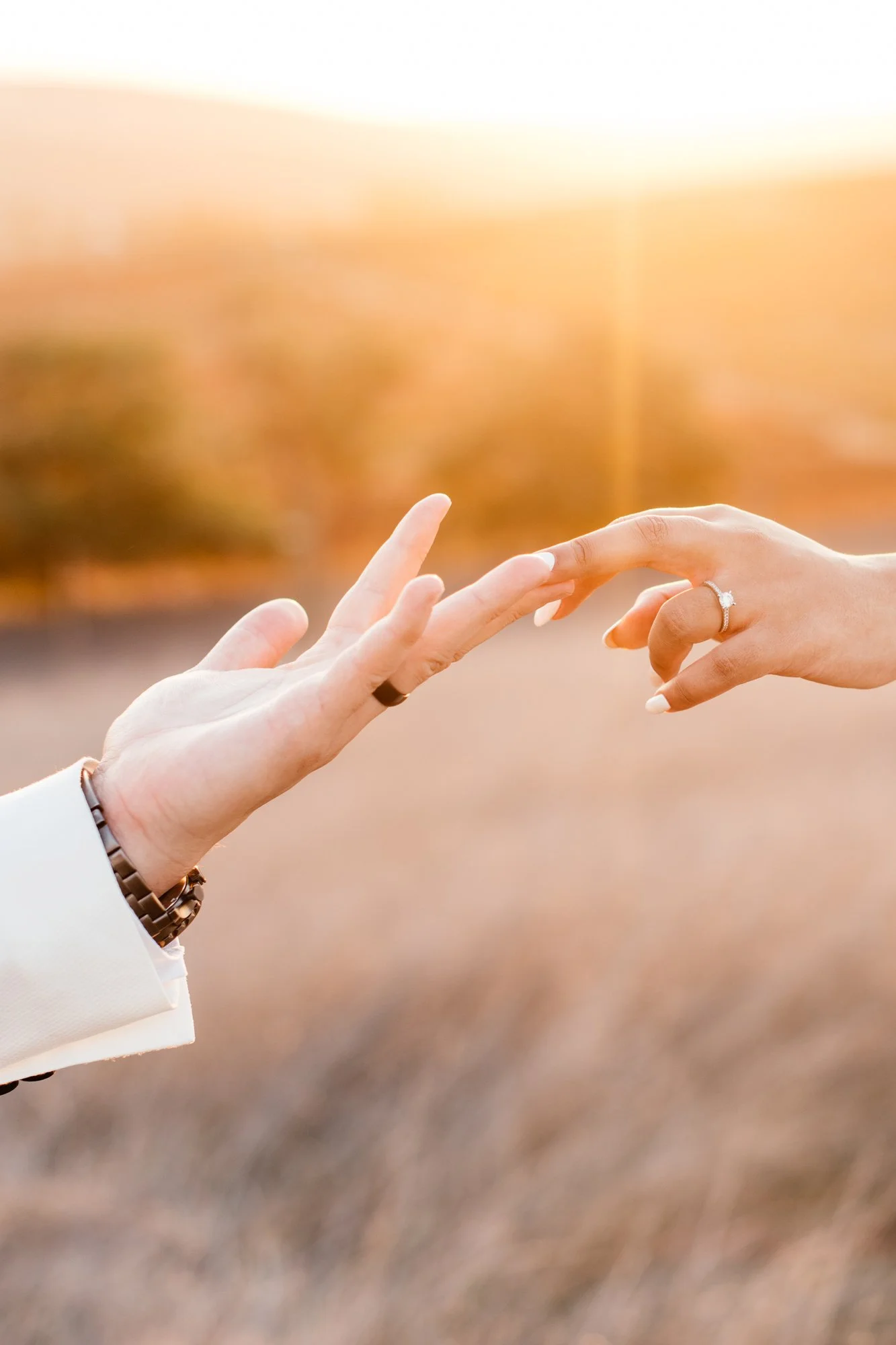 Two hands reaching towards each other, one with an engagement ring, in a sunset setting.