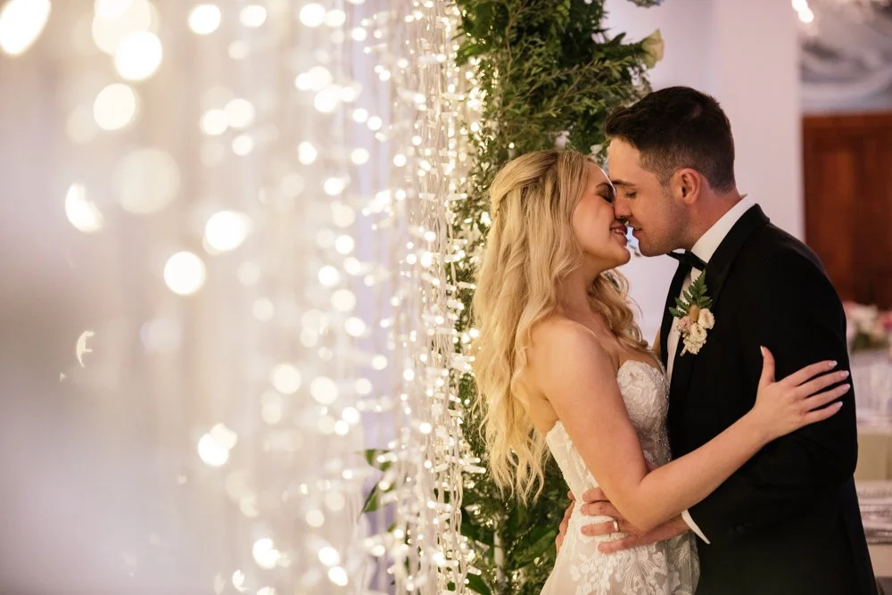 A bride and groom are close together, touching foreheads and smiling, during their wedding reception, with decorative lights and greenery in the background.