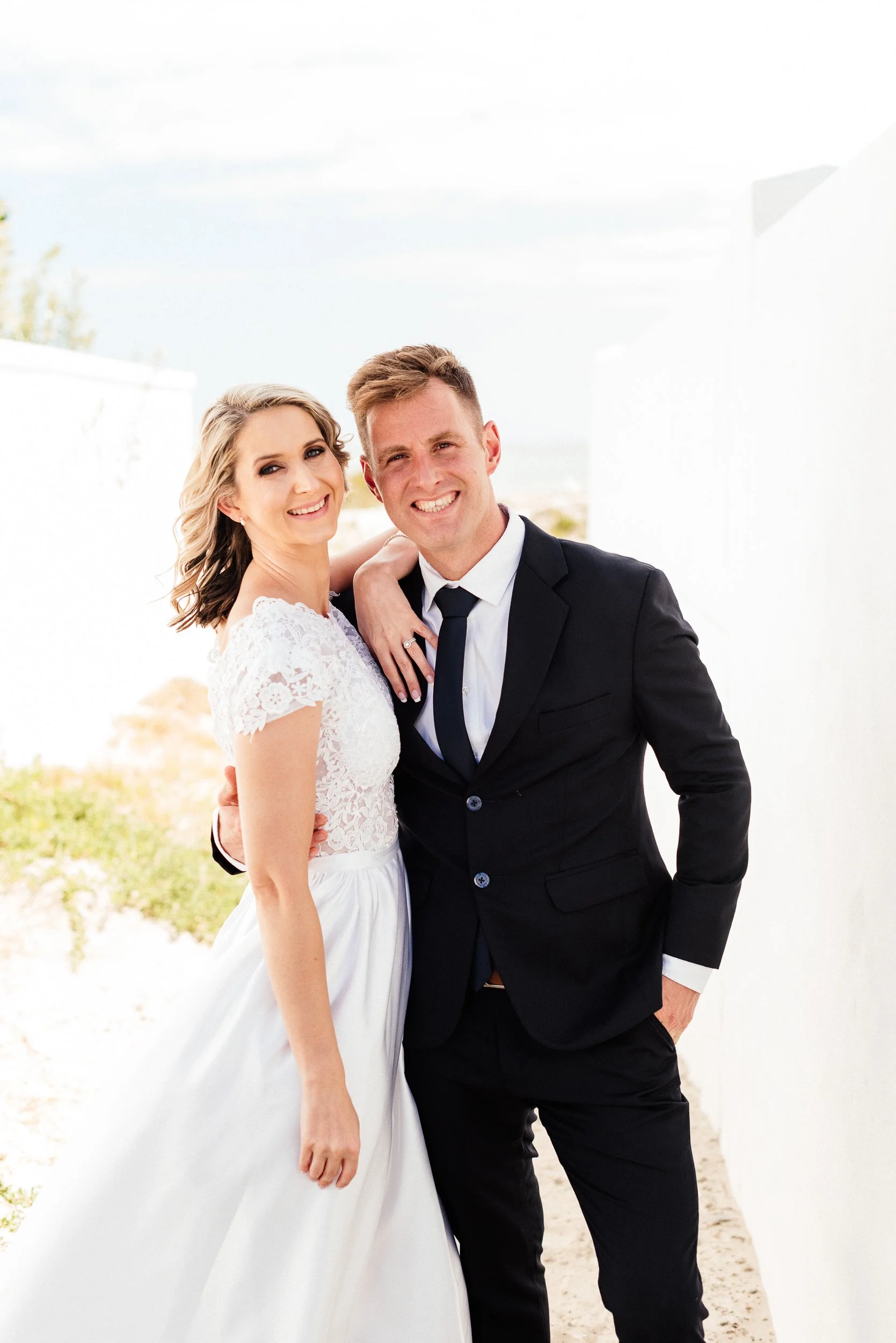 A smiling bride and groom standing together outdoors, with the groom in a black suit and the bride in a white wedding dress, against a bright sky and white architectural structure.