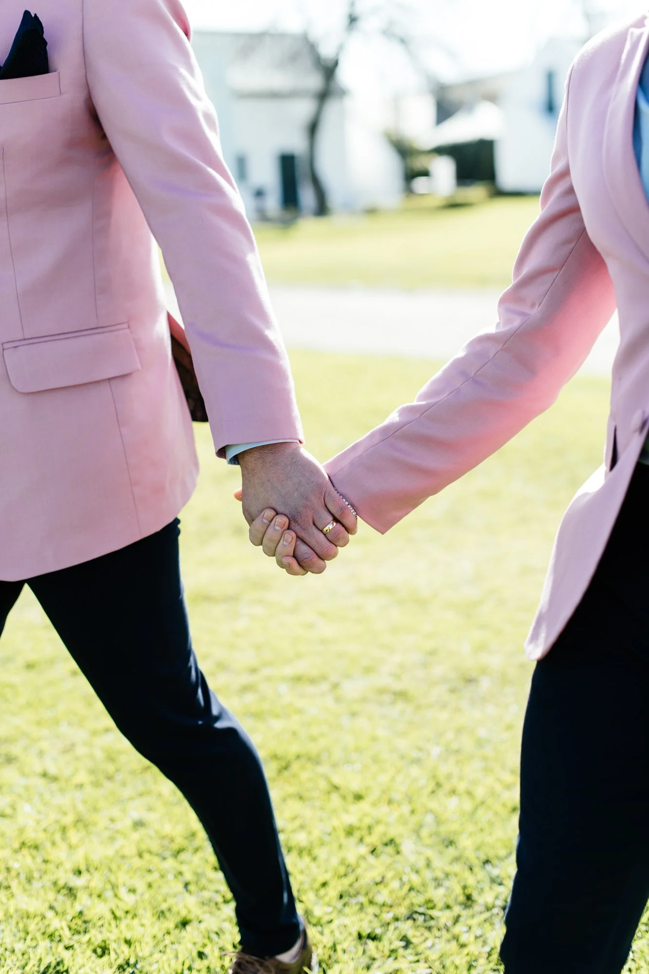 Two grooms in pink blazers holding hands outdoors in sunlight, with houses and trees in the background.  Love wins.