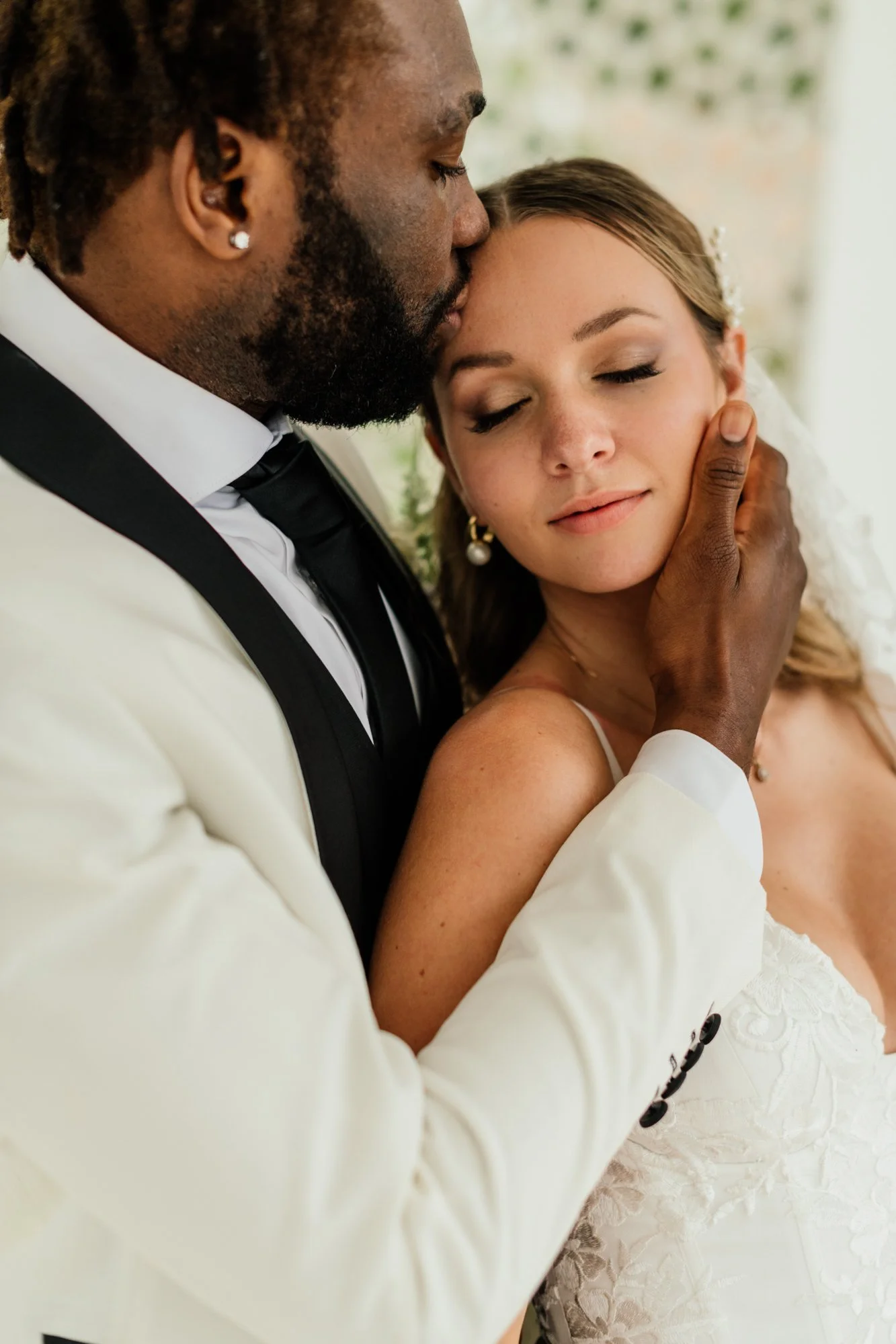 The groom with a beard and earrings gently kissing the forehead of his bride, closed eyes, and pearl earrings, during a wedding moment.