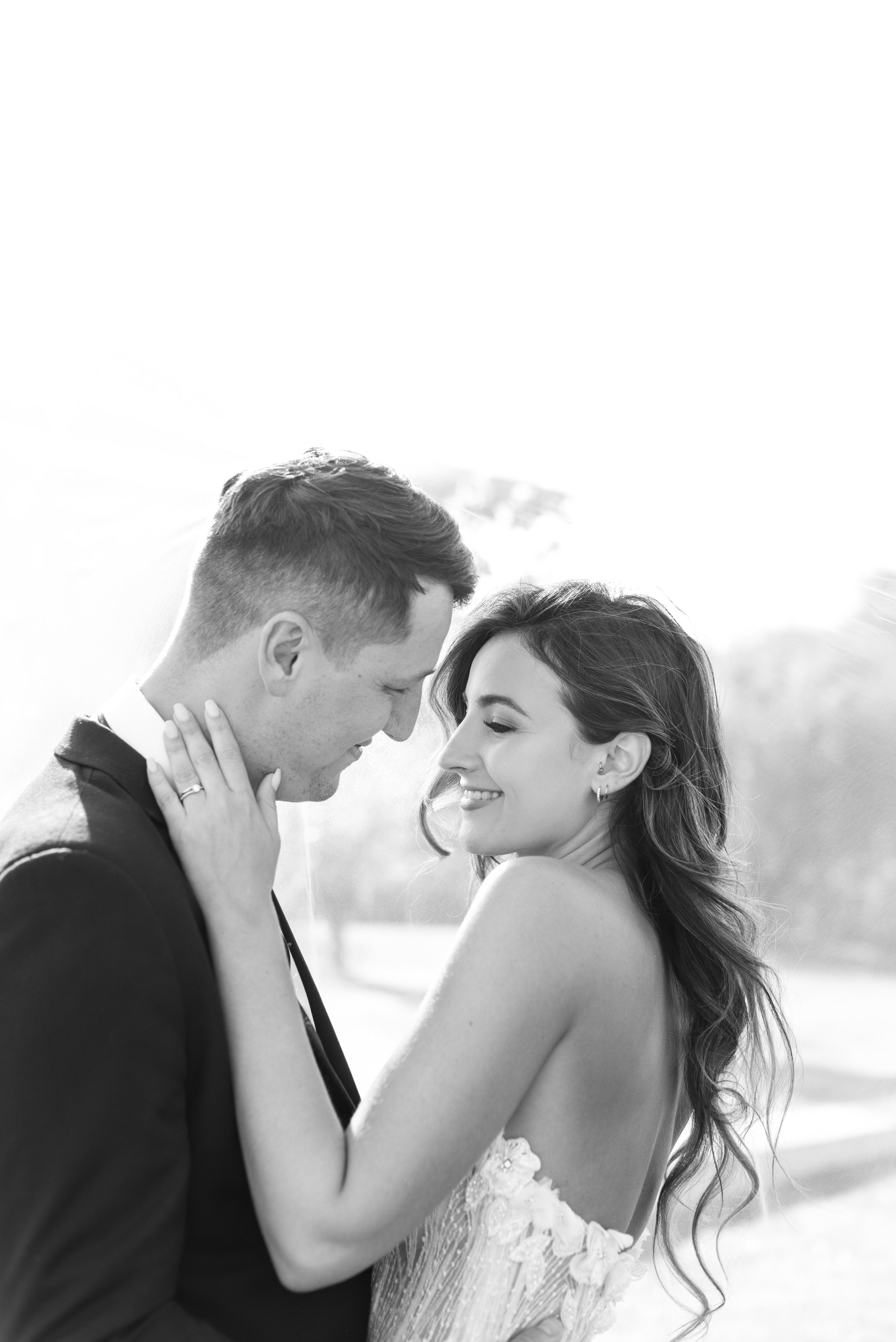 A black and white photo of a couple on their wedding day, standing close with foreheads touching, the groom's hand on the bride's neck, both smiling with closed eyes.