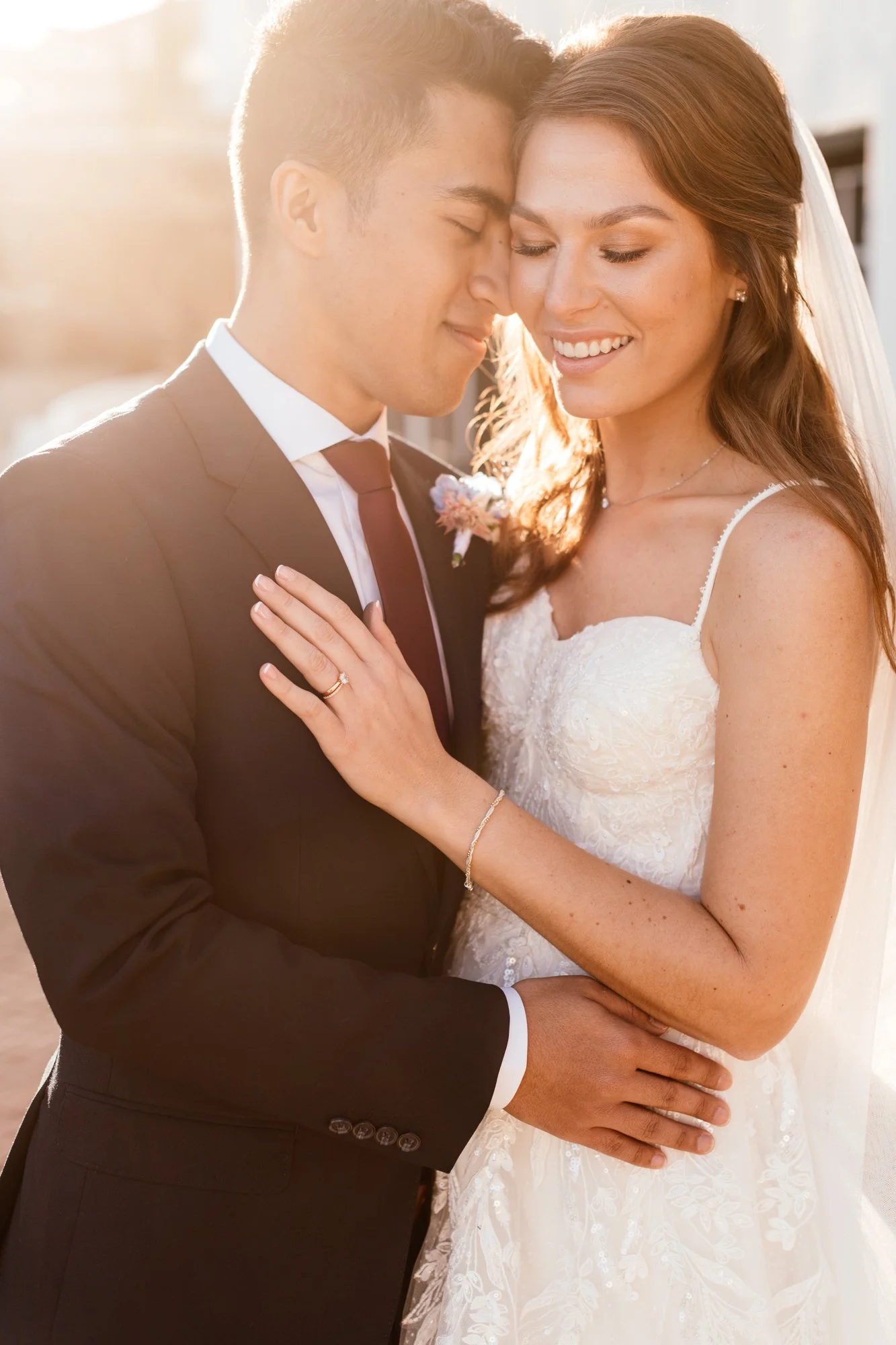 A newlywed couple sharing a moment with their foreheads touching, standing outdoors during sunset. The groom is dressed in a tuxedo, and the bride is wearing a white wedding gown with a veil.