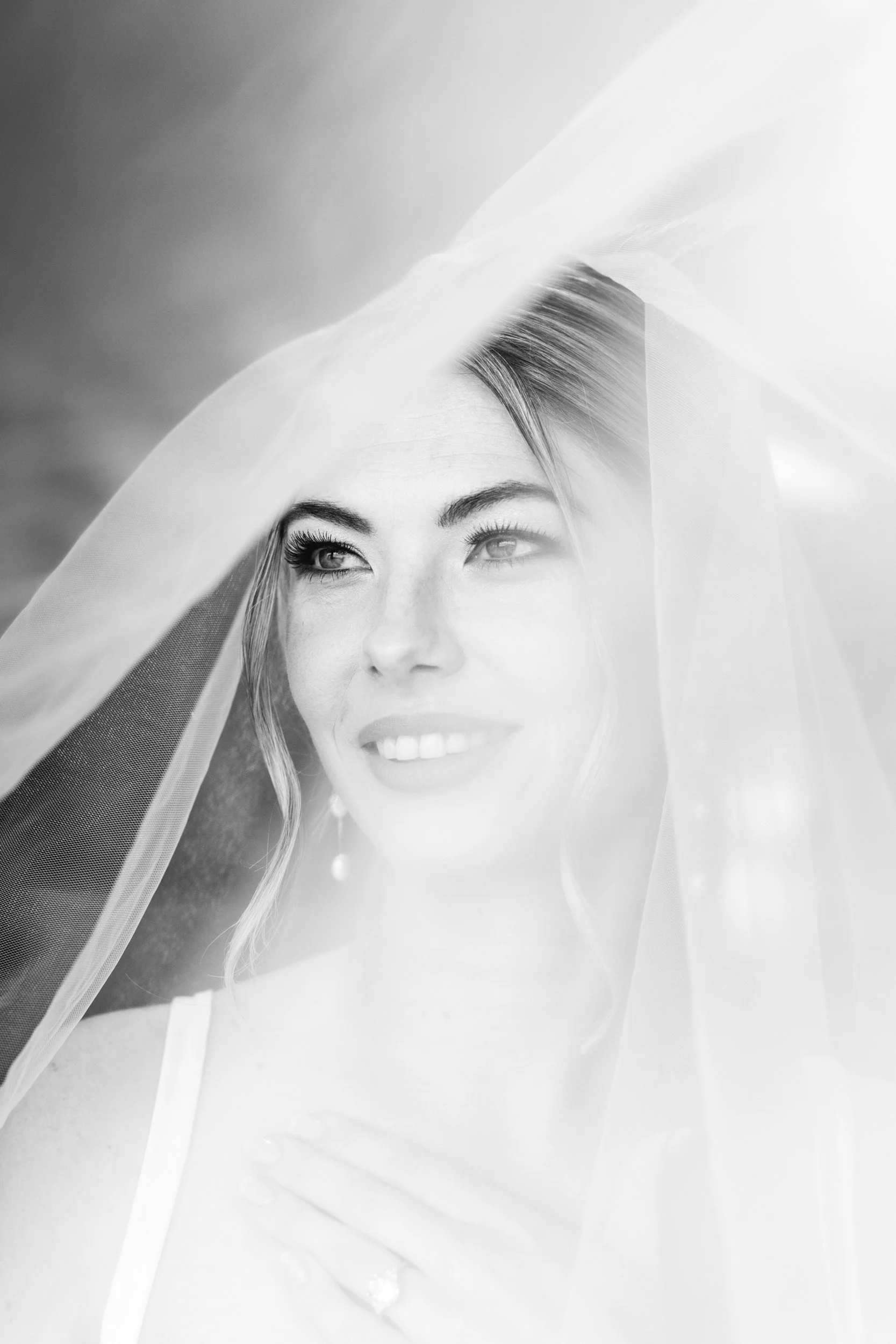 Black and white close-up of a woman with fair skin and blonde hair, smiling with her eyes looking slightly upward. She is wearing a veil.
