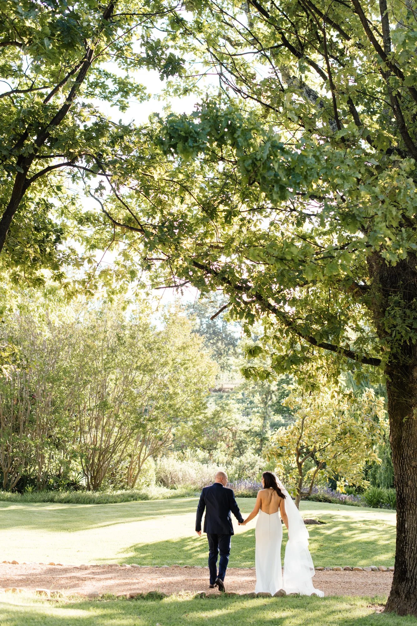 A bride and groom walking hand in hand outdoors on a sunny day, surrounded by large trees and lush greenery.
