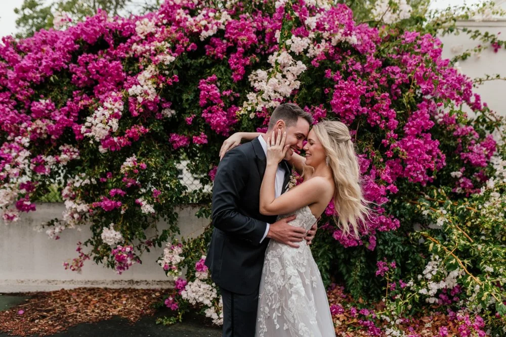 A newlywed couple in wedding attire sharing an intimate moment in front of a vibrant pink and white flowering bush.