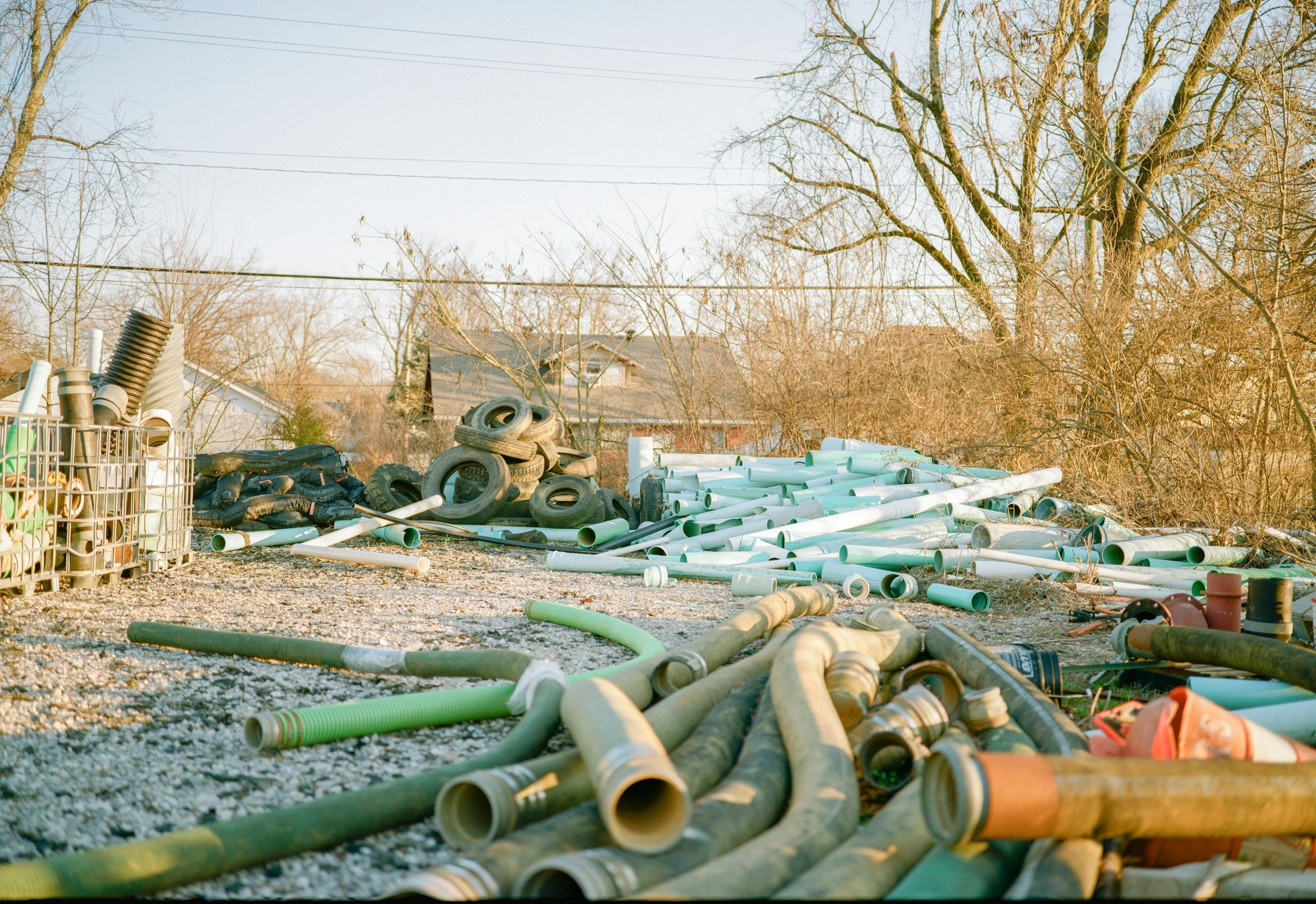 Yard filled with discarded pipes, tires, and various debris with bare trees and houses in the background.