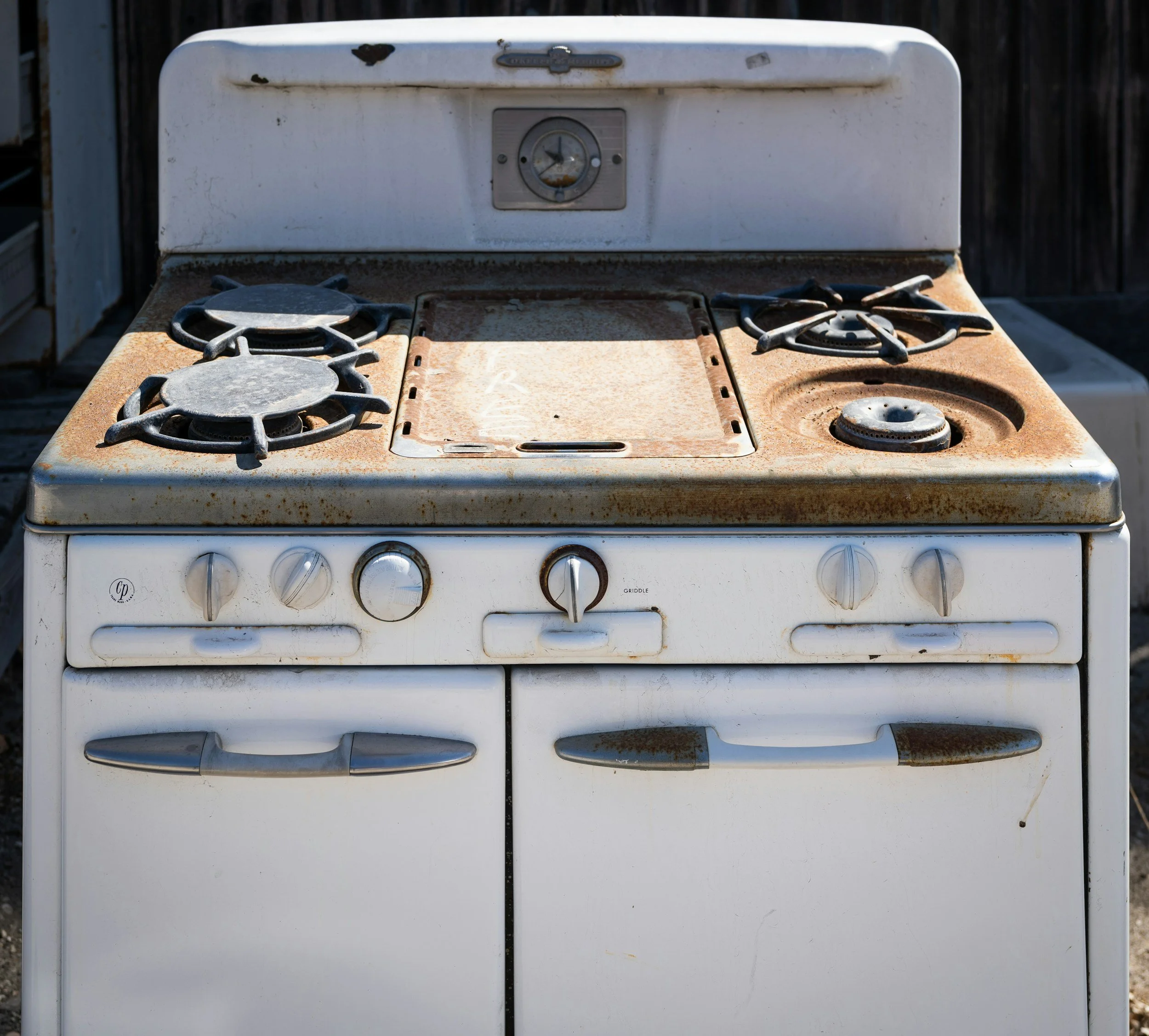 An old, rusty white gas stove with three burners and a control panel, located outdoors with a wooden fence in the background.