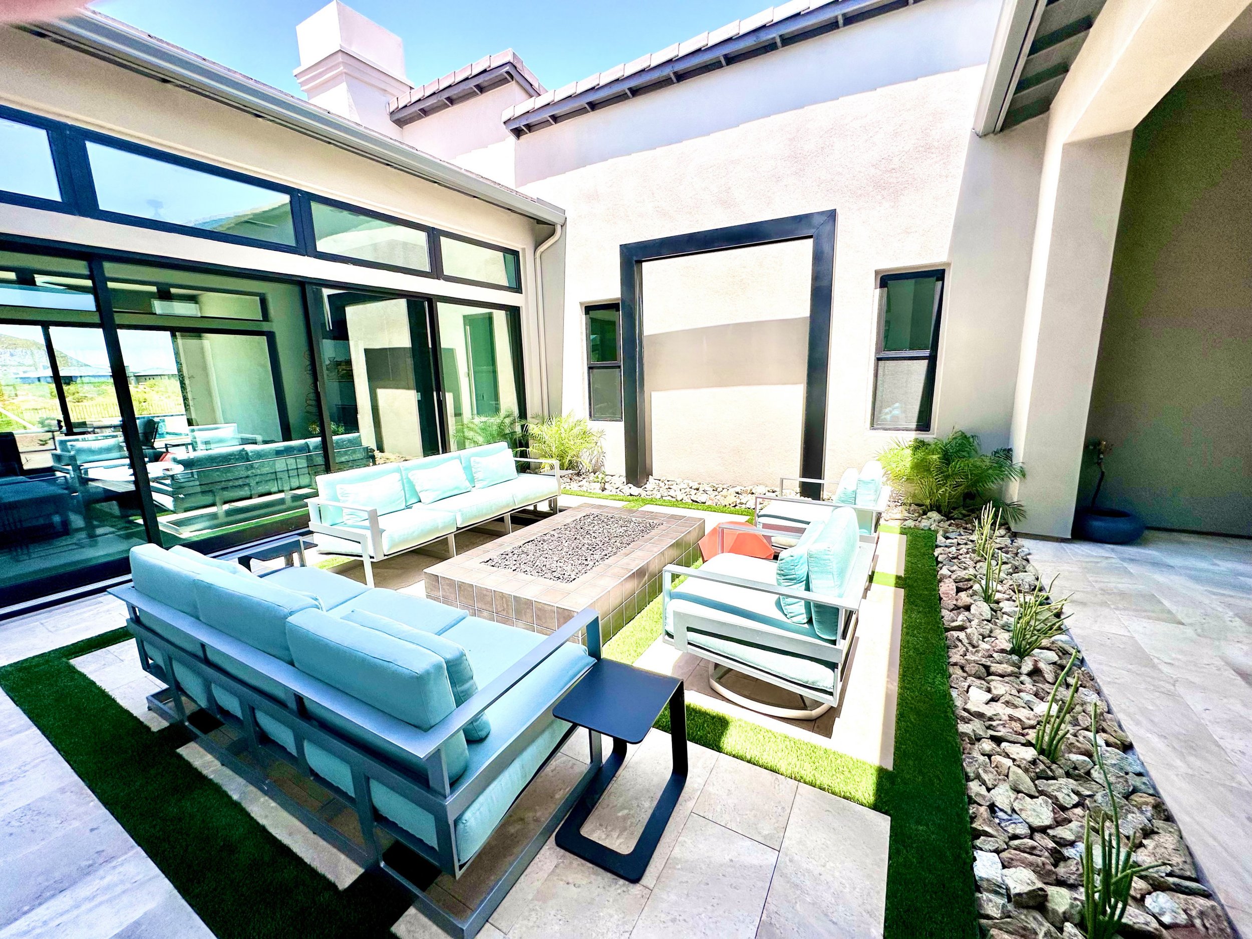 An outdoor patio area with modern furniture, including light blue sofas, armchairs, and black side tables, surrounded by rocks and potted plants, with a glass wall and a house with a chimney in the background.