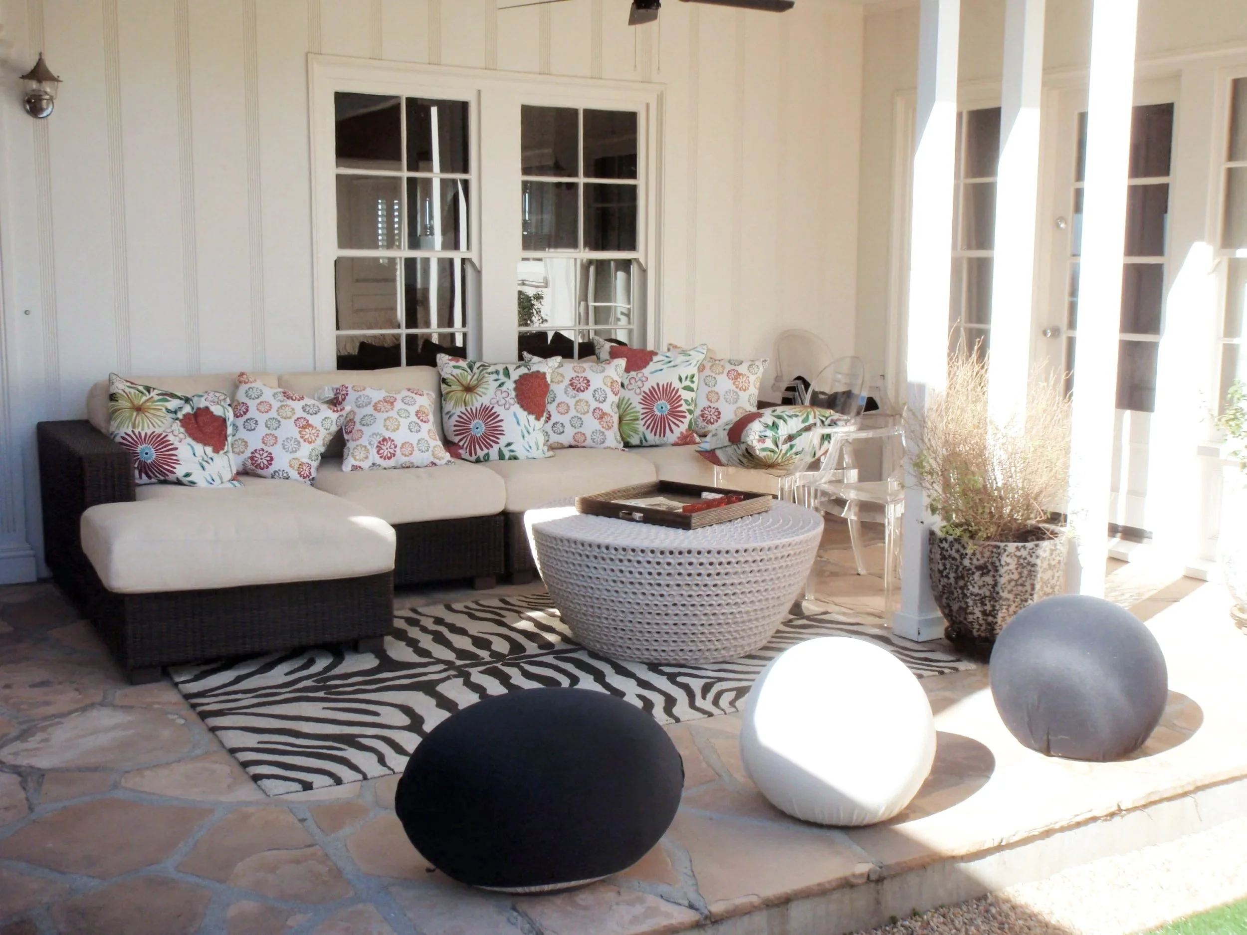 A cozy living room with a sectional sofa adorned with floral-patterned pillows, a round wicker coffee table, zebra print rug, and large windows with white curtains allowing sunlight to brighten the space. There are decorative elements including a potted plant and hanging light fixtures.