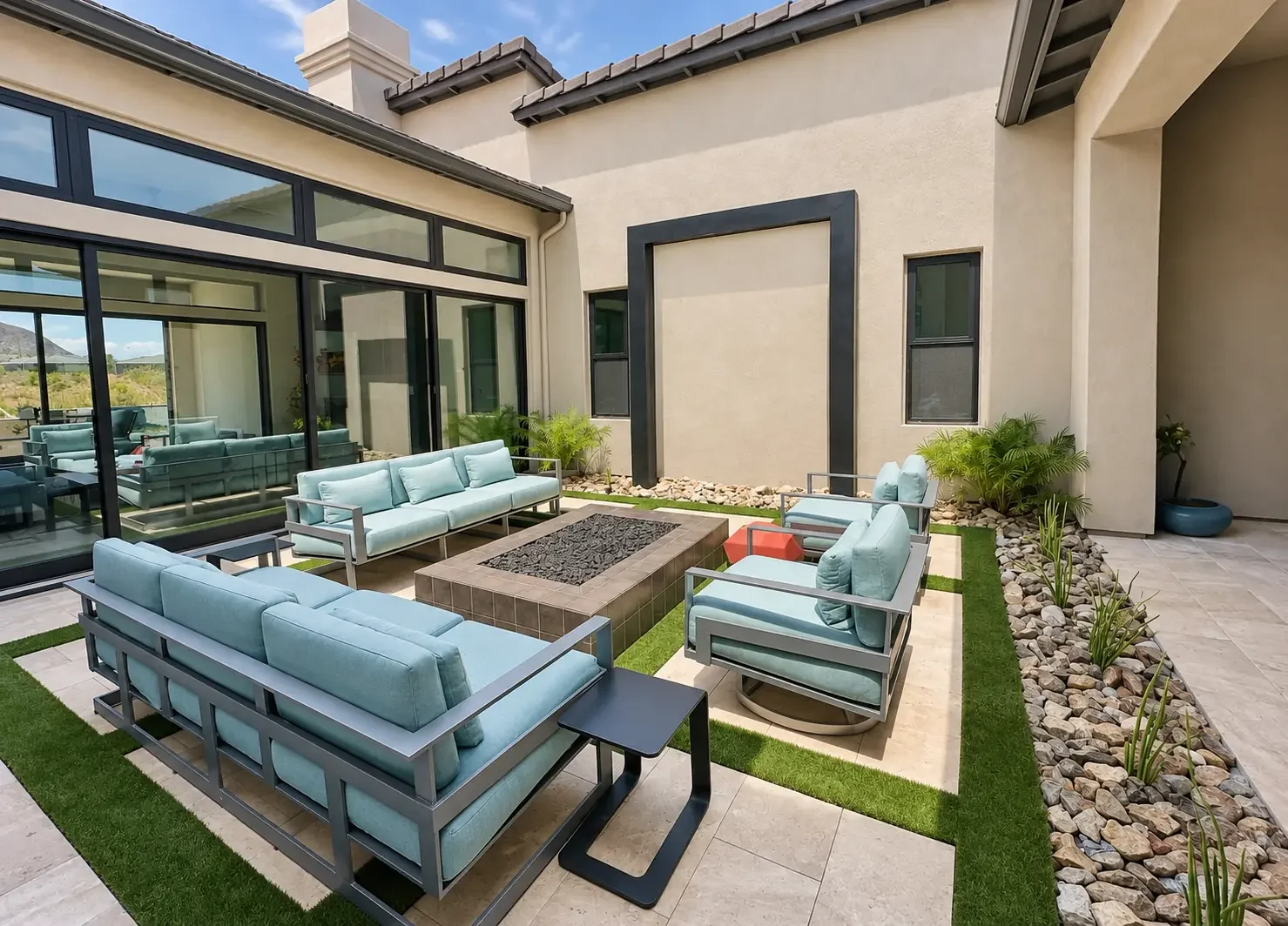 An outdoor patio area with modern furniture, including light blue sofas, armchairs, and black side tables, surrounded by rocks and potted plants, with a glass wall and a house with a chimney in the background.