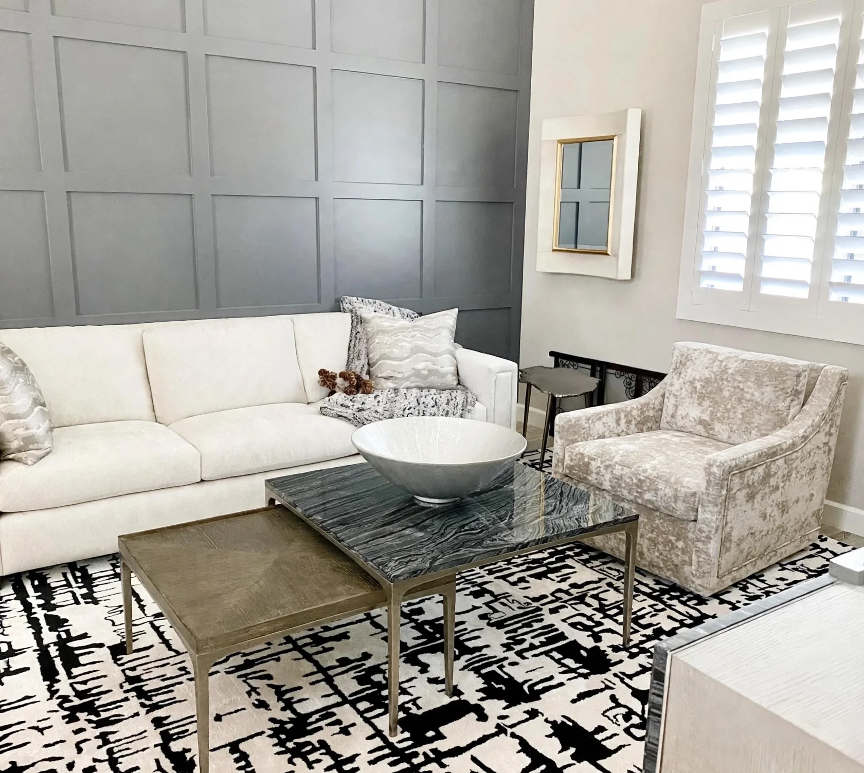 Living room with a modern television mounted on the wall, a marble and metal console table, a patterned black and white rug, a beige sofa, a glass-topped coffee table, a standing lamp, and window blinds.
