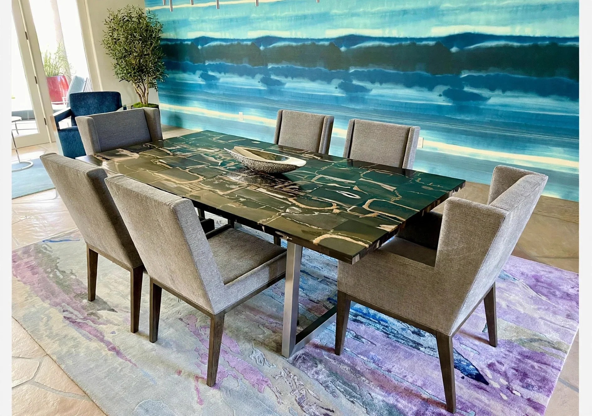 A dining room with a modern black marble table surrounded by eight light grey upholstered chairs. The room features a large, abstract blue wall mural, a potted plant, a glass door, and a colorful area rug.