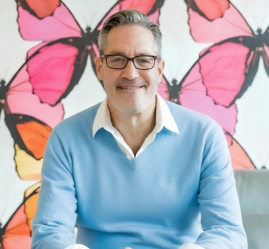 A man with gray hair, glasses, and a beard, smiling and sitting in front of a colorful butterfly wall mural.