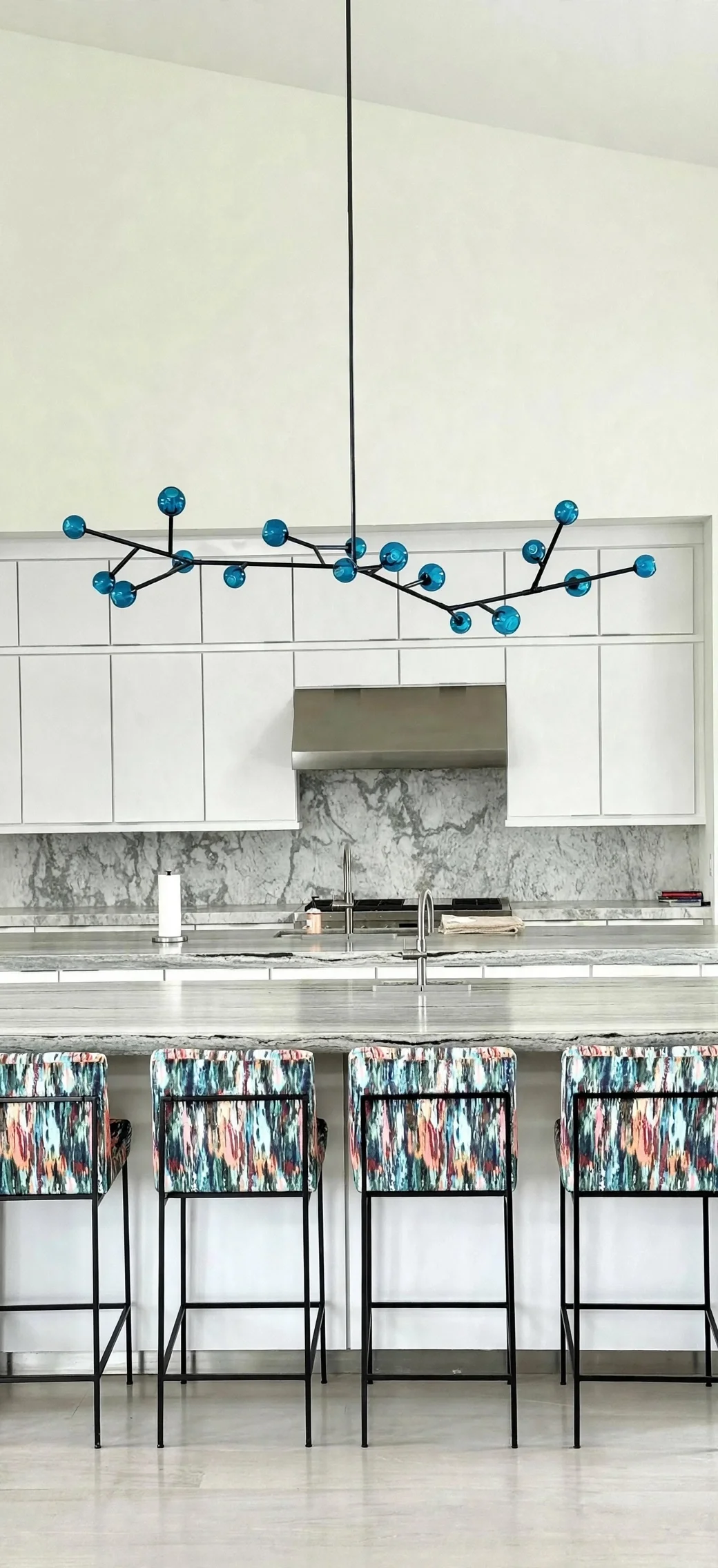 Modern kitchen with white cabinets, marble backsplash, stainless steel range hood, and colorful patterned bar stools at a marble countertop, with a blue geometric chandelier hanging above.