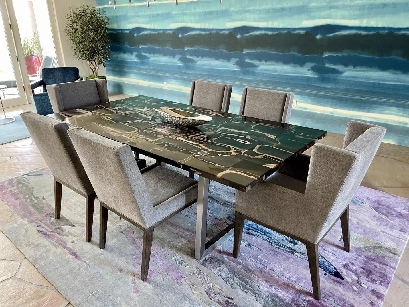 A dining room with a modern black marble table surrounded by eight light grey upholstered chairs. The room features a large, abstract blue wall mural, a potted plant, a glass door, and a colorful area rug.
