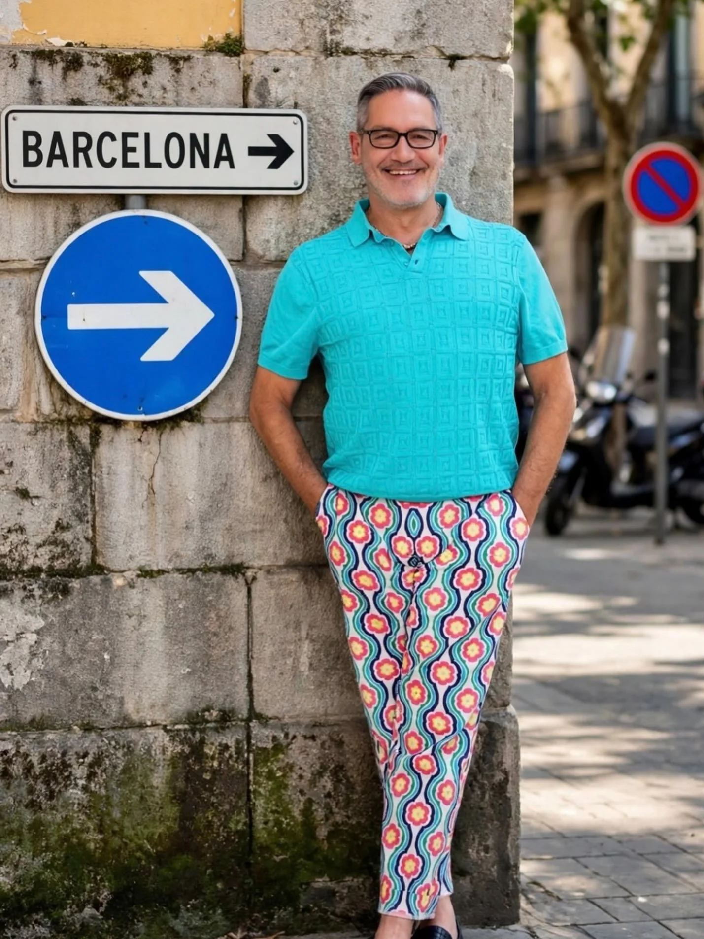 A smiling man wearing glasses, a turquoise shirt, colorful patterned pants, and dark shoes, standing next to a stone wall with road signs, in an urban setting with trees and buildings in the background.