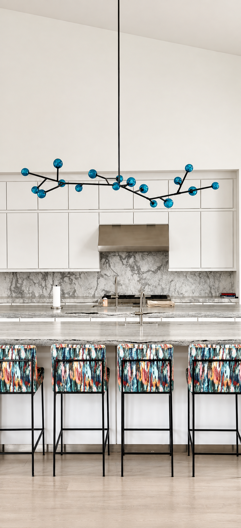 Modern kitchen with white cabinets, marble backsplash, stainless steel range hood, and colorful patterned bar stools at a marble countertop, with a blue geometric chandelier hanging above.