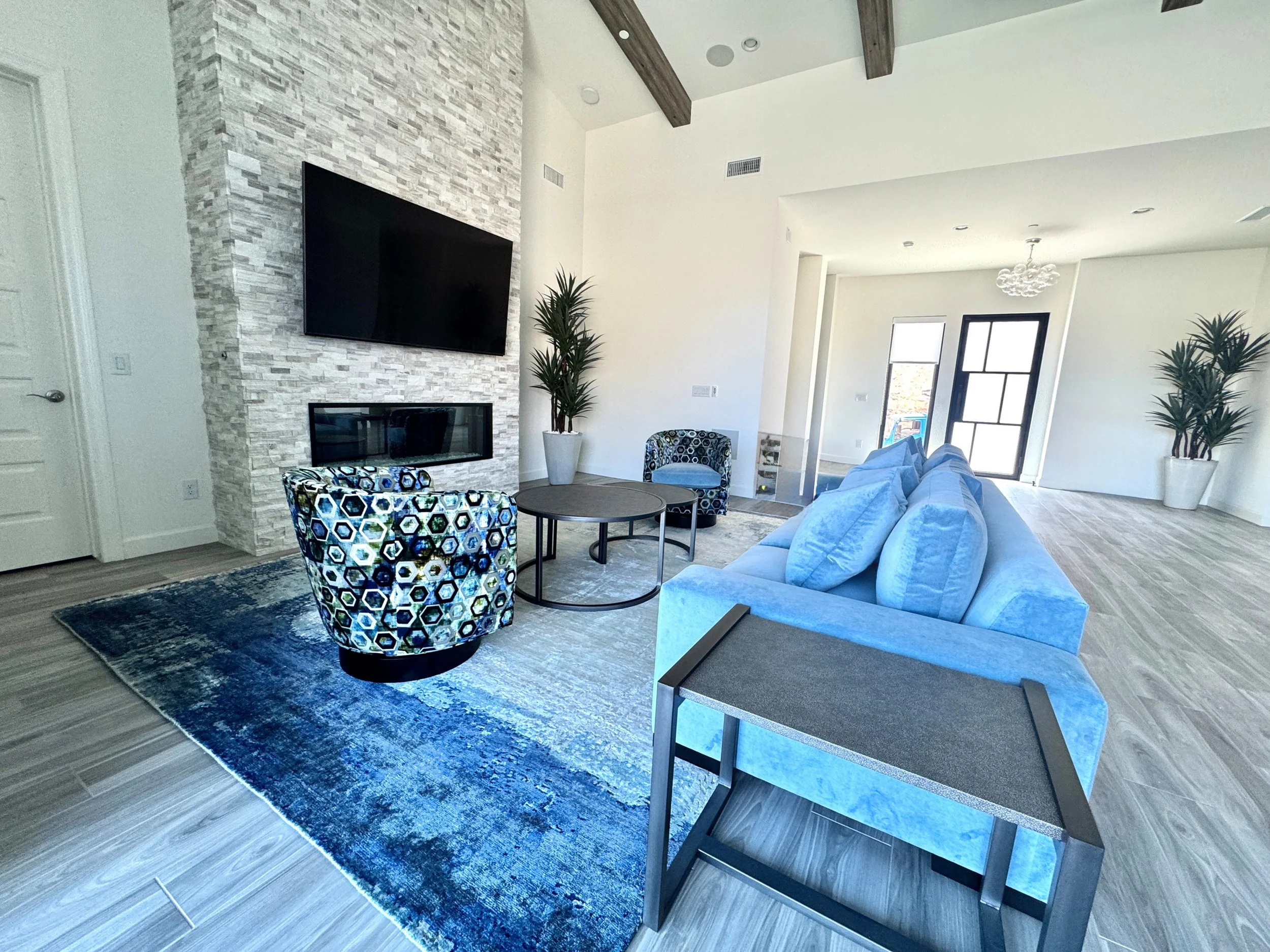 Modern living room with white walls, a stone accent wall featuring a mounted flat-screen TV and a fireplace, a blue sofa, a patterned armchair, a round coffee table, and large windows with black frames letting in natural light. There are two tall potted plants and a blue area rug.