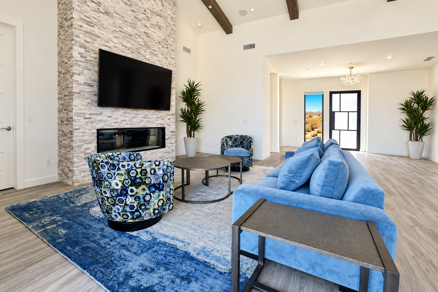 Modern living room with white walls, a stone accent wall featuring a mounted flat-screen TV and a fireplace, a blue sofa, a patterned armchair, a round coffee table, and large windows with black frames letting in natural light. There are two tall potted plants and a blue area rug.