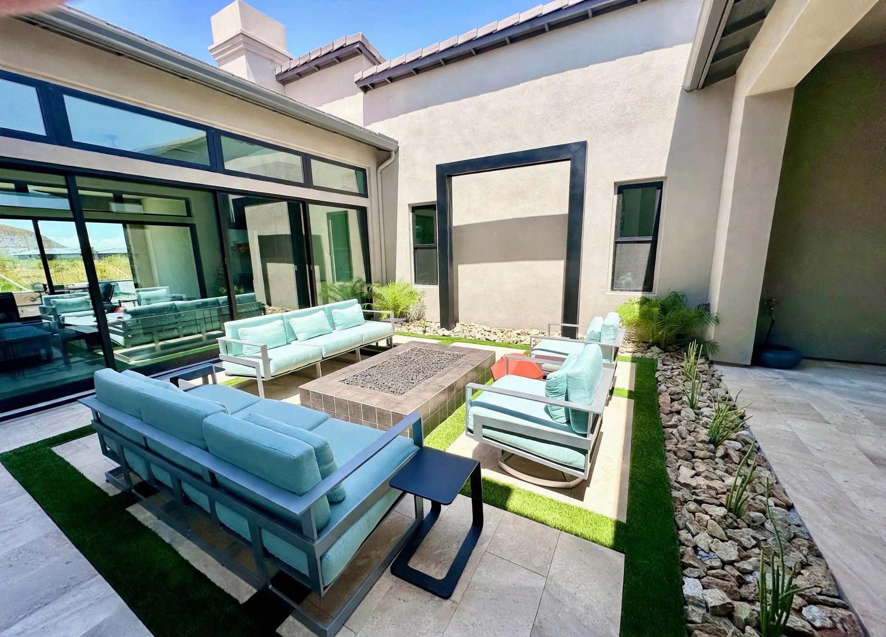 An outdoor patio area with modern furniture, including light blue sofas, armchairs, and black side tables, surrounded by rocks and potted plants, with a glass wall and a house with a chimney in the background.